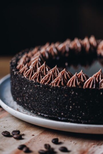 A whole chilled mocha cheesecake is sitting on a pink plate, that is on a wooden board with coffee beans scattered over it. The cheesecake has a dark crust and chocolate piped cream on top, in the centre is a coffee syrup.