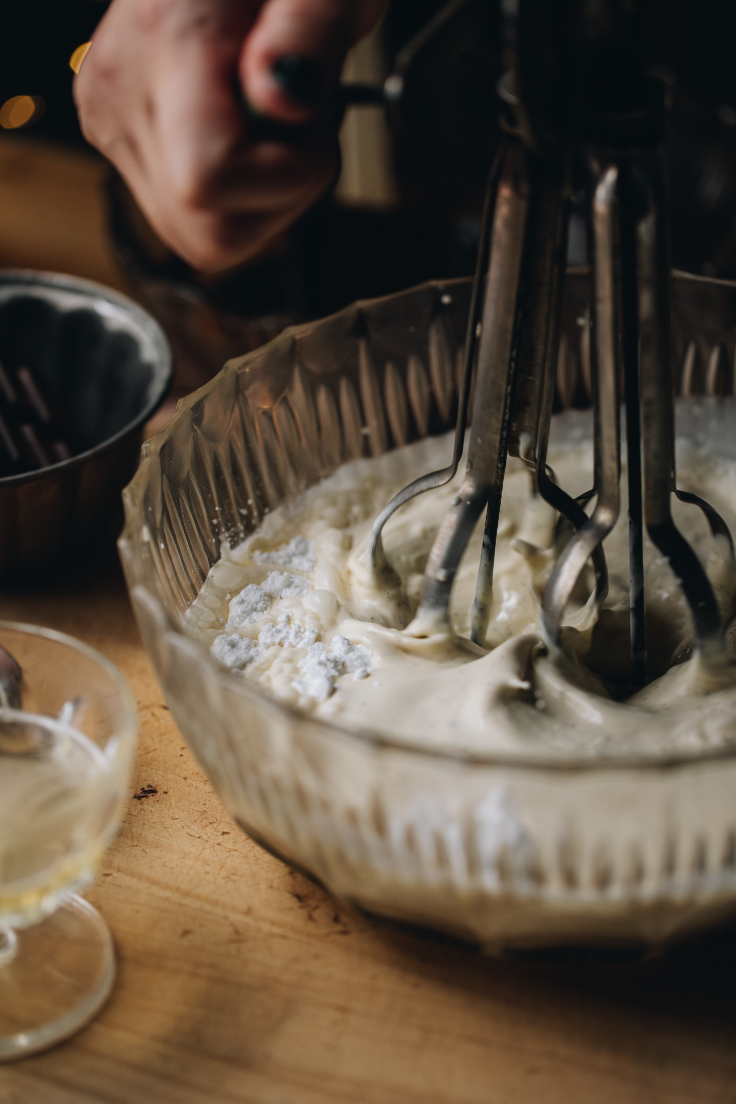 A close shot of cream being whipped in a vintage glass bowl. It is being whipped by a vintage egg beater on a wooden table. 