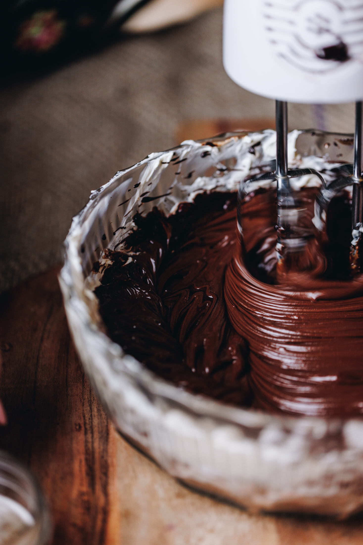 The chocolate cheesecake filling of the Chilled Mocha Cheesecake is being whipped with a white hand mixer. It is in a glass vintage bowl on a wooden table. 