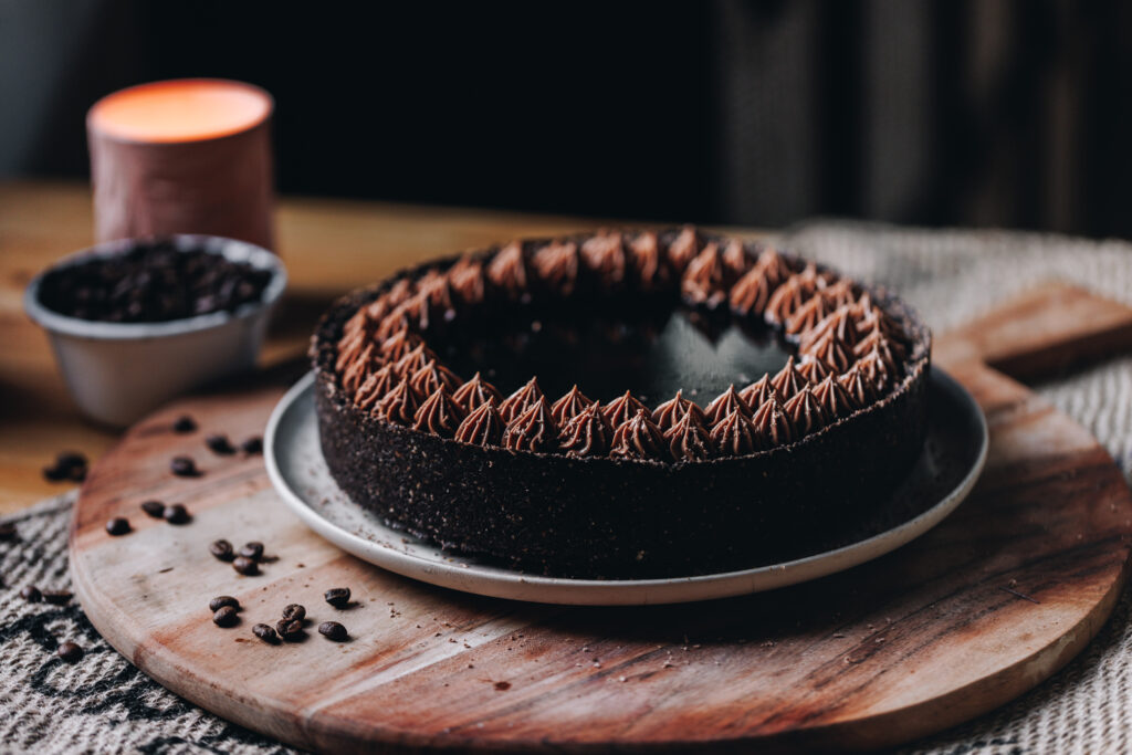 A landscape photo shows a whole chilled mocha cheesecake is sitting on a pink plate, that is on a wooden board with coffee beans scattered over it. The cheesecake has a dark crust and chocolate piped cream on top, in the centre is a coffee syrup.