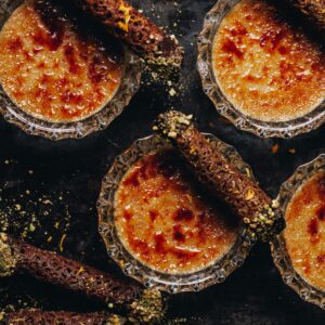 On a black tray is a flat lay of freshly baked chai creme brûlée, the caramelised sugar on top is seen along with mini brandy snaps sitting on top of each dessert.