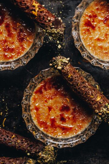 On a black tray is a flat lay of freshly baked chai creme brûlée, the caramelised sugar on top is seen along with mini brandy snaps sitting on top of each dessert.
