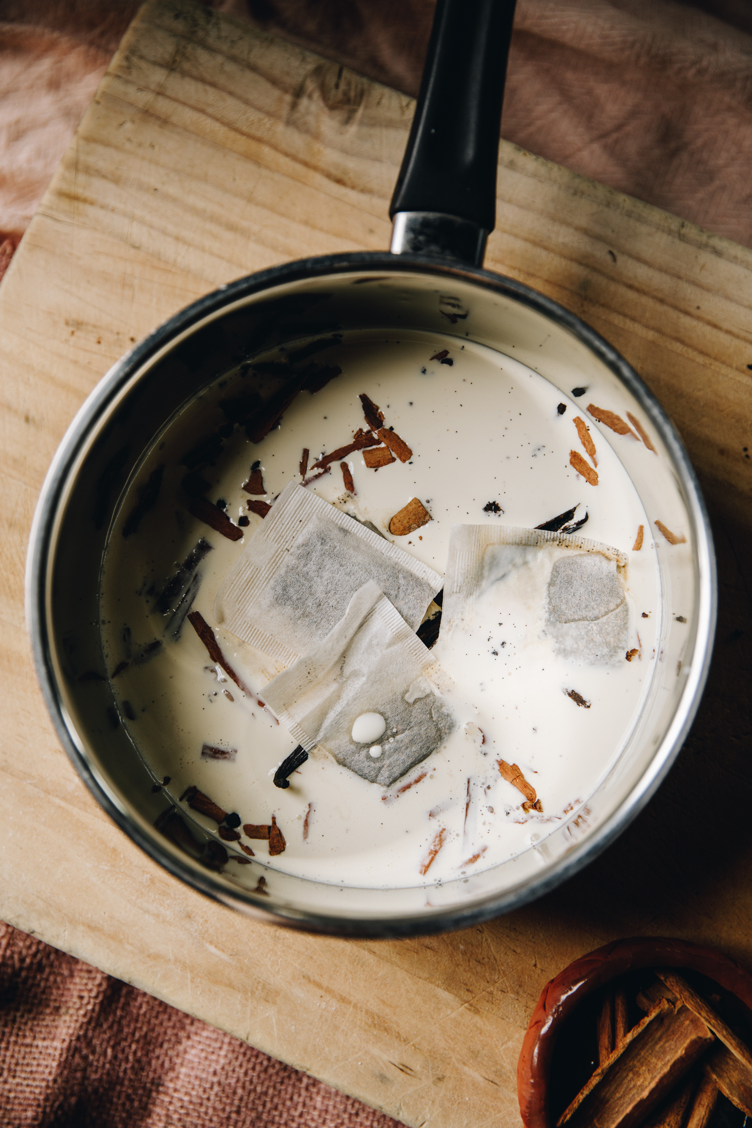 A flat lay shot shows a pot with chai tea bags, cinnamon sticks and cream in it. It is sitting on a wooden board.