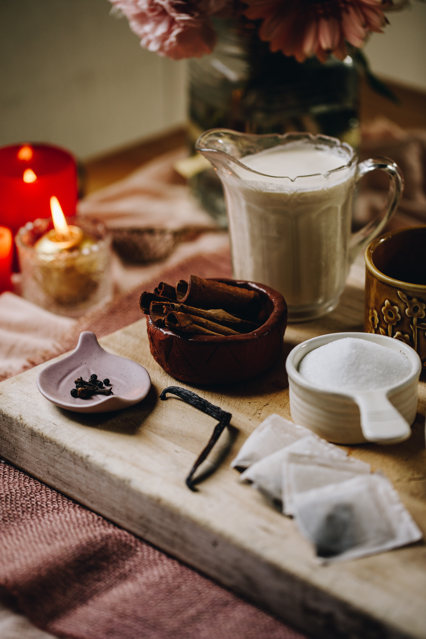 Chai Créme Brûlée spice ingredients are in different vintage bowls and are sitting on a wooden board with flowers and a candle in the background.