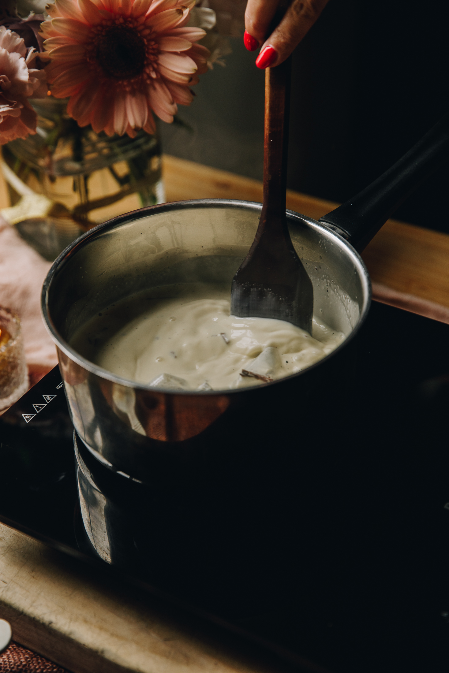 A pot with chai tea bags, cinnamon sticks and cream in it is being stirred on a black portable stove top. There is flowers in the background, it is all on a wooden table. 