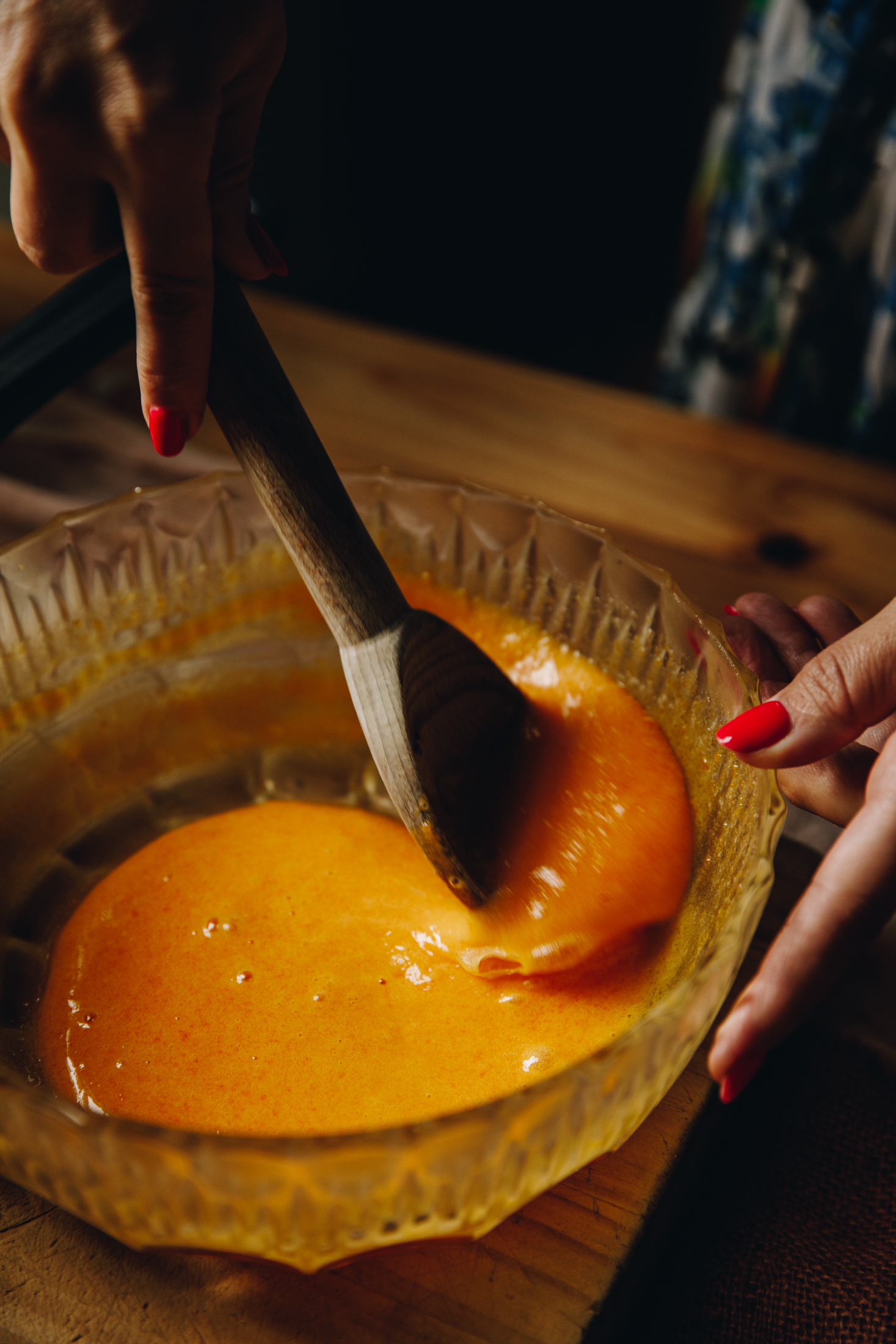 In a glass vintage bowl egg yolks are being stirred with a wooden spoon. The bowl is on a wooden table. 