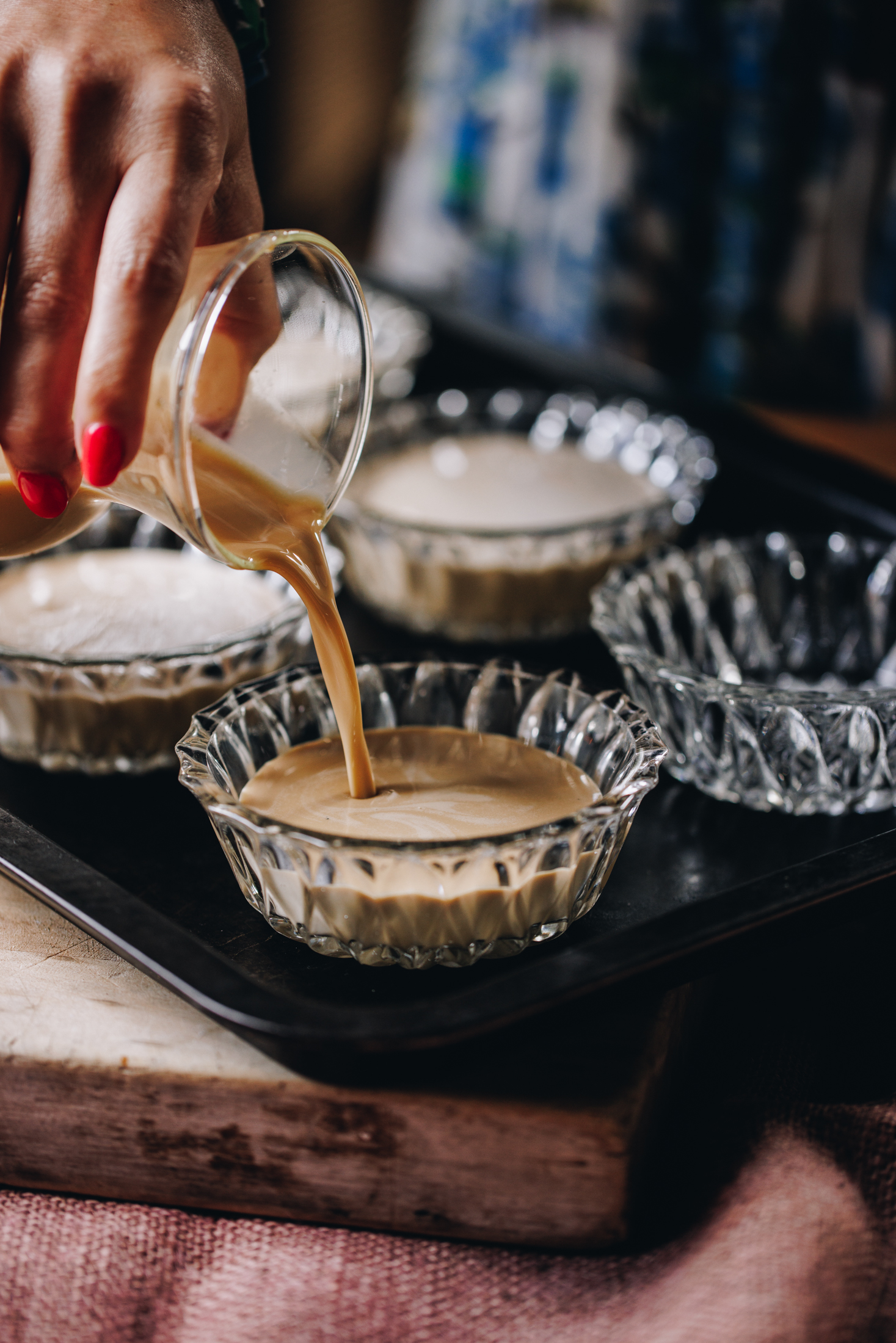 Glass ramekins are on a black tray, the chai creme brûlée mixture is being poured in to one of the ramekins. The tray is on a wooden board on a pink tablecloth. 