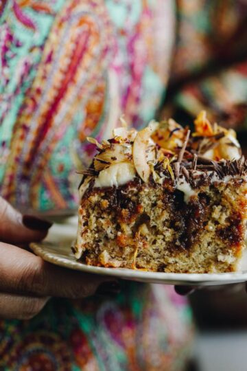 On a vintage plate sits a piece of a banana and caramel poke cake that reveals the gooey caramel that has been poked inside the cake. On top of grated chocolate and cream cheese frosting.