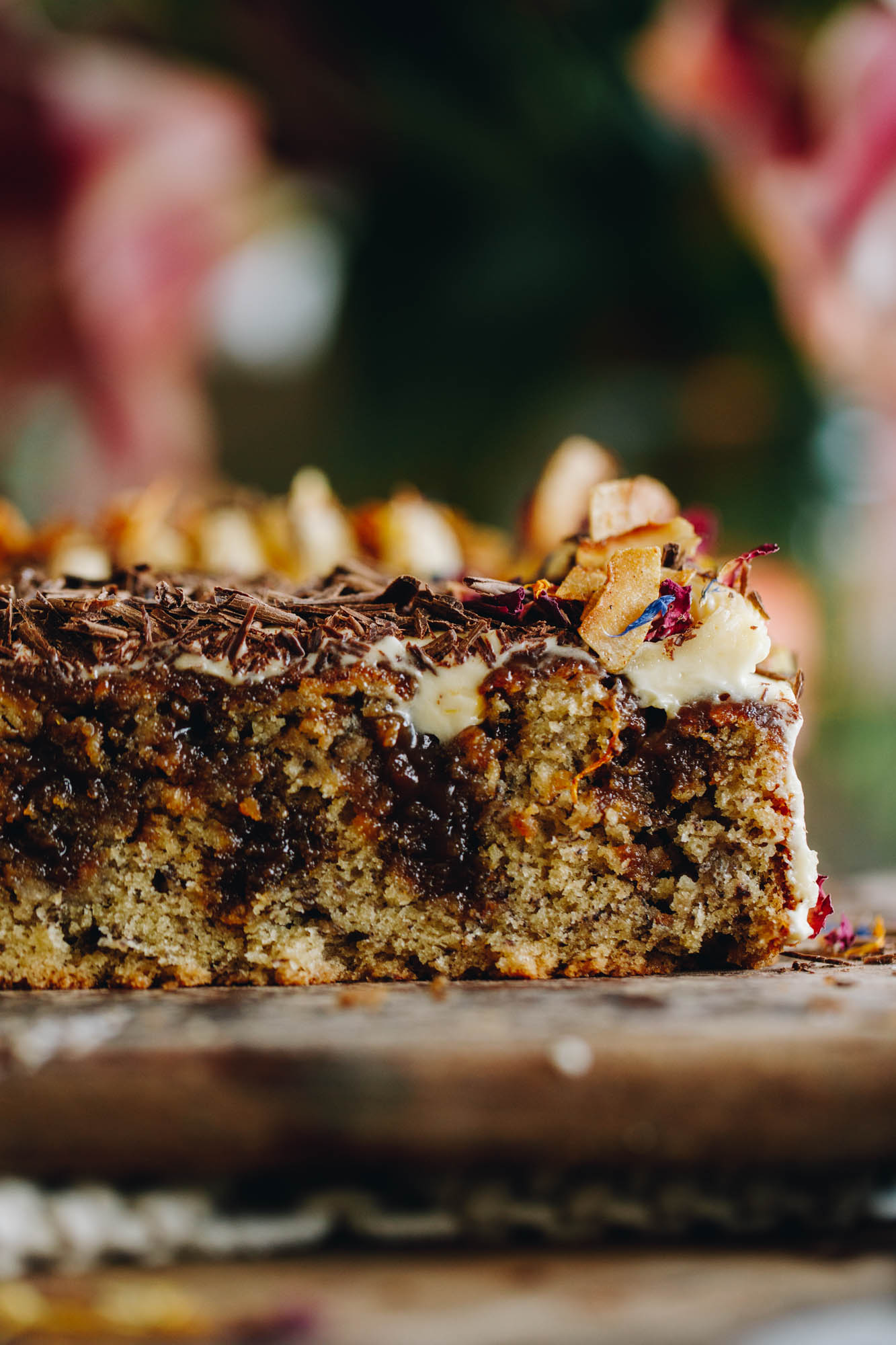 On a wooden board sits a banana and caramel poke cake that has been cut in half and reveals the gooey caramel that has been poked inside the cake. On top of grated chocolate, cream cheese frosting and coconut chips.