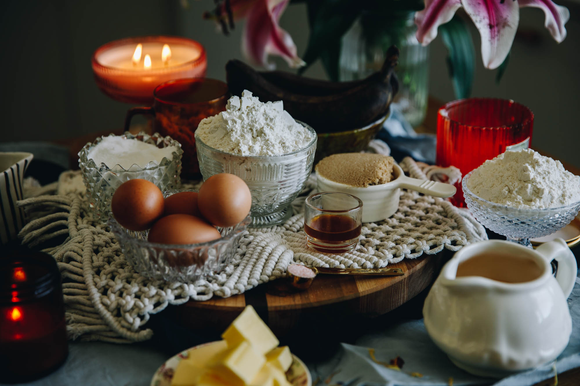 Cake ingredients are in different vintage bowls and are sitting on a wooden board on top of wooden table. There is pink flowers and a candle burning in the background.