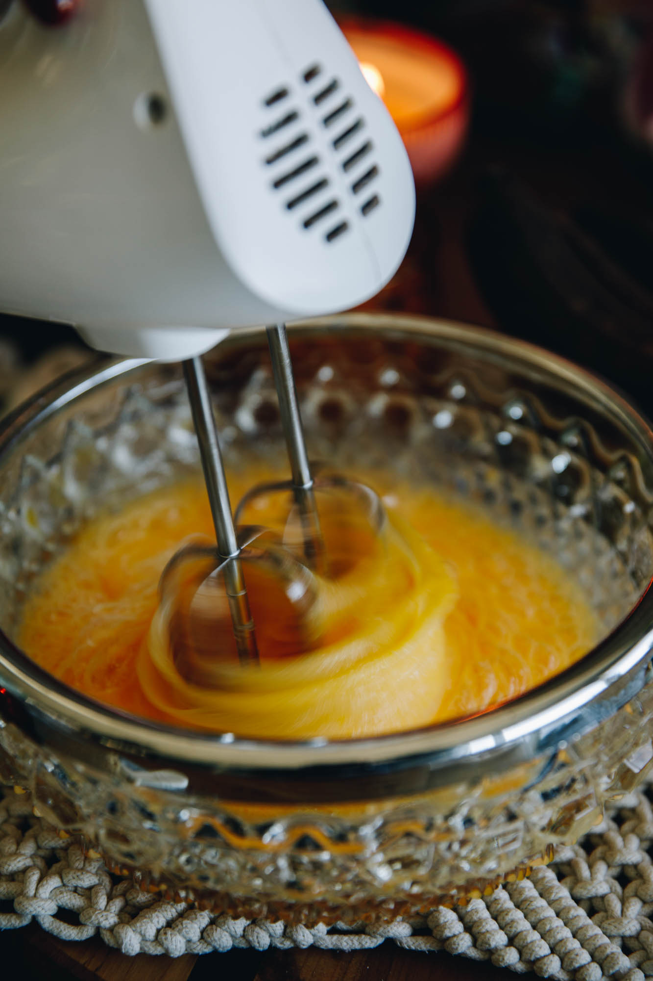 In a vintage glass bowl is eggsand sugar being whipped with a w white hand mixer. It is sitting on a cream crocheted mat.
