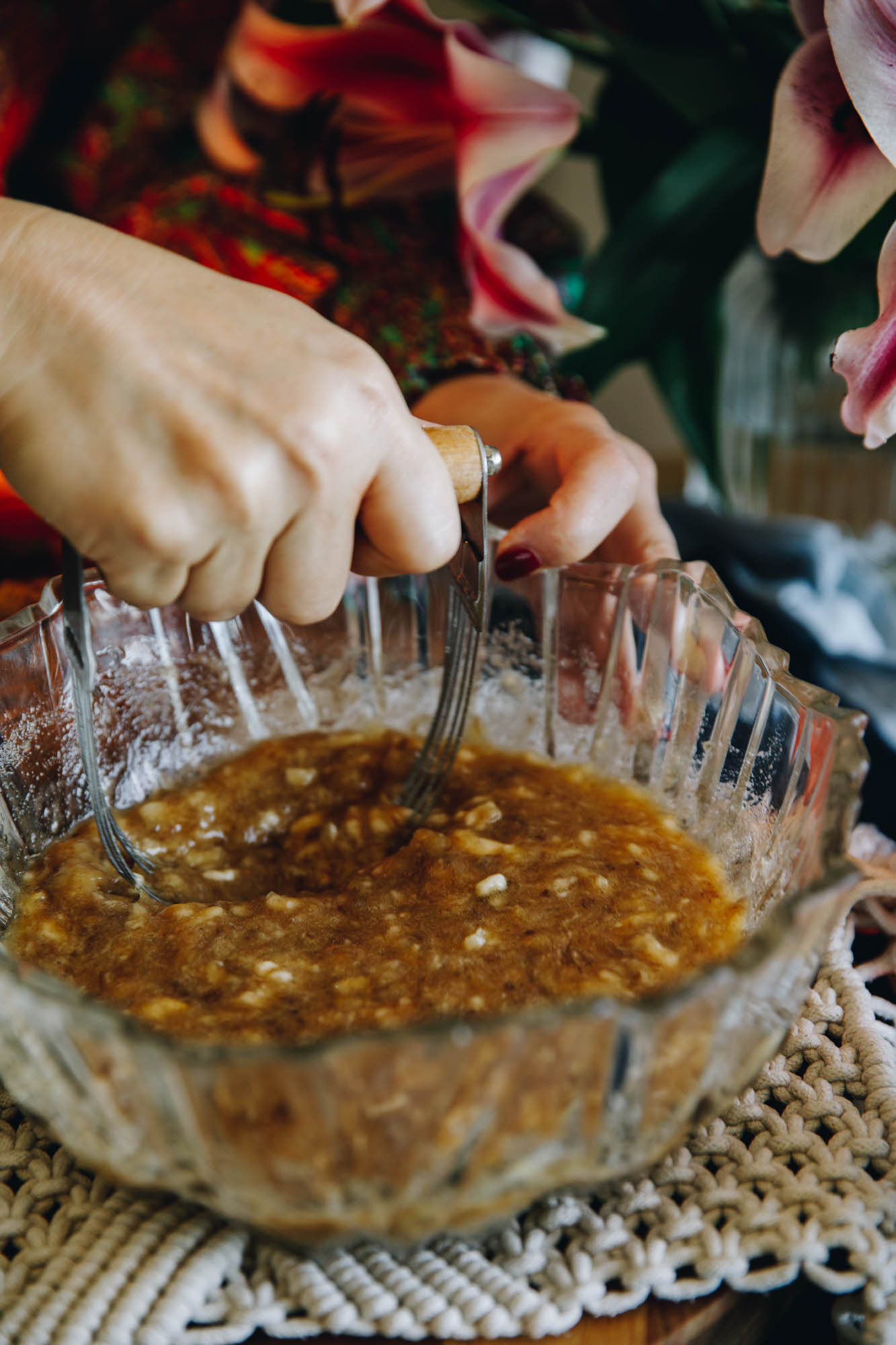 In a vintage glass bowl is mashed banana being mashed with a vintage pastry cutter. It is sitting on a cream crocheted mat.