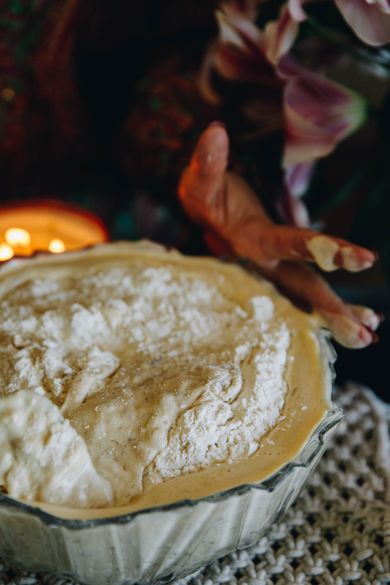 In a vintage glass bowl is the egg and banana mixture of banana cake. It is filled to the brim and is being folded with a spatula. It is sitting on a cream crocheted mat.