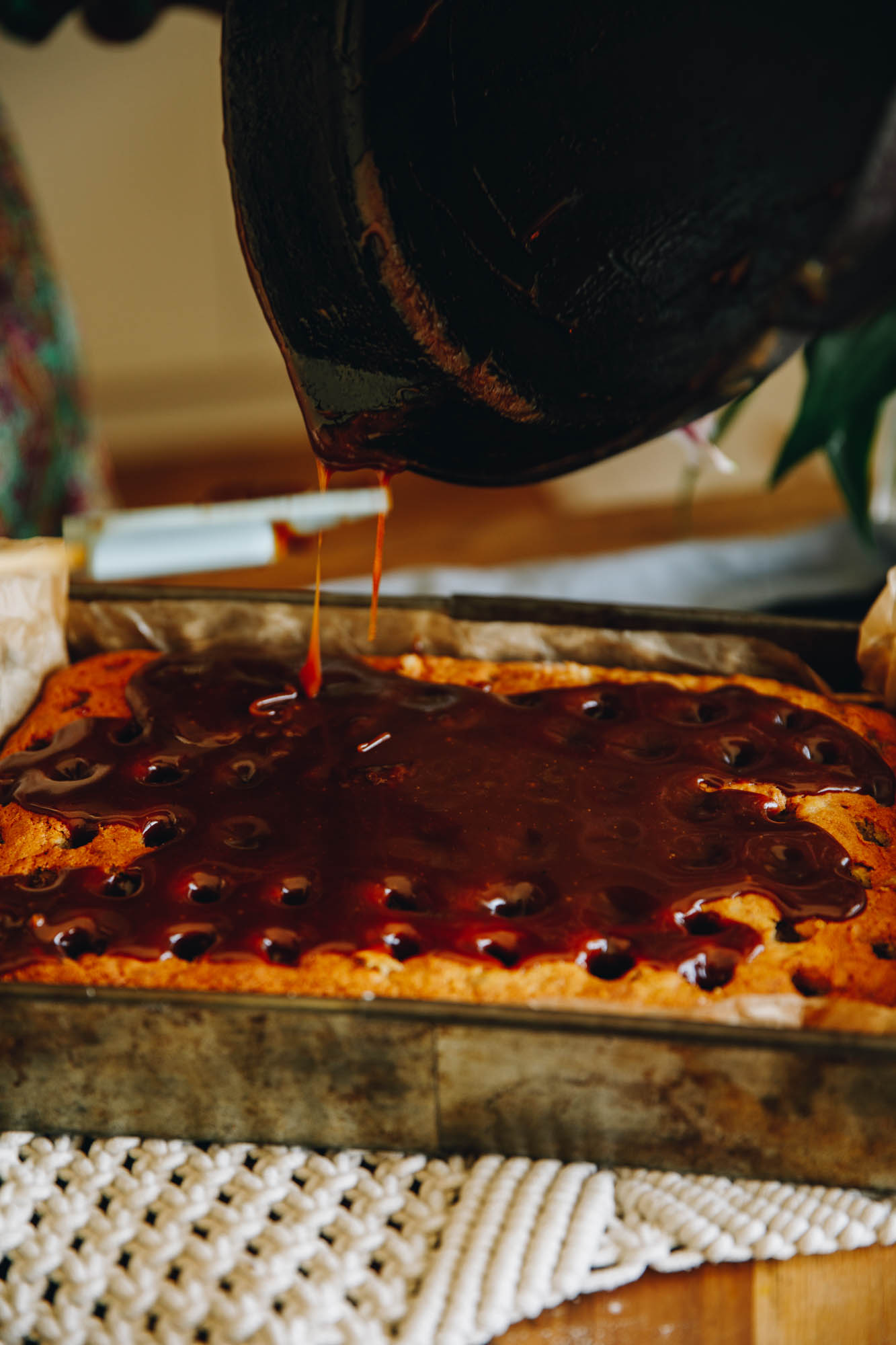 On a cream crocheted mat sits a freshly baked banana cake that has holes all over the top of it. Deep coloured caramel has been poured on top and the drips from a black cast iron pot are in view.
