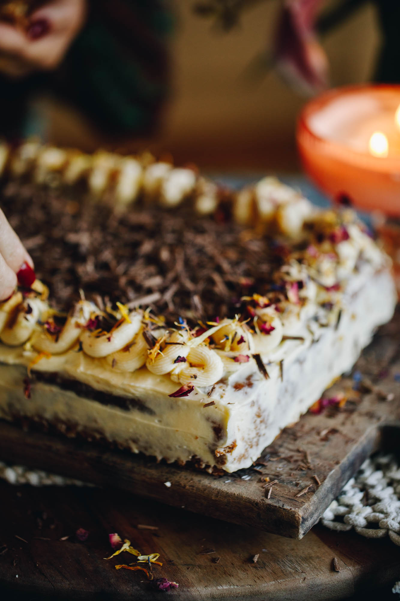 A freshly baked banana and caramel poke cake has been decorated with cream cheese frosting with swirls on top of it with small flower petals and coconut chips. Chocolate has been grated in the centre of the slab cake.