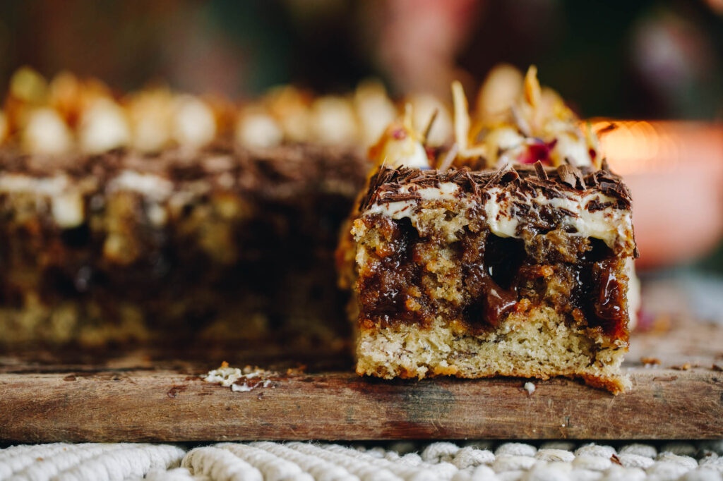 A landscape shot shows a baked banana and caramel poke cake slice with oozy caramel coming out of it, it is on a wooden board.