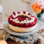 A whole peanut butter jelly cheesecake sits on a wooden table with natural fabrics under it and flowers in the background. The cheesecake layers show the biscuit base, a peanut butter filling, topped with a raspberry jelly with whole raspberries in it. The top has been decorated with piped cream, freeze-dried raspberries and flower petals.