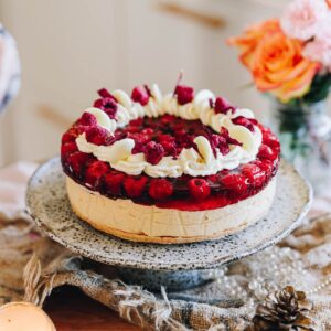 A whole peanut butter jelly cheesecake sits on a wooden table with natural fabrics under it and flowers in the background. The cheesecake layers show the biscuit base, a peanut butter filling, topped with a raspberry jelly with whole raspberries in it. The top has been decorated with piped cream, freeze-dried raspberries and flower petals.