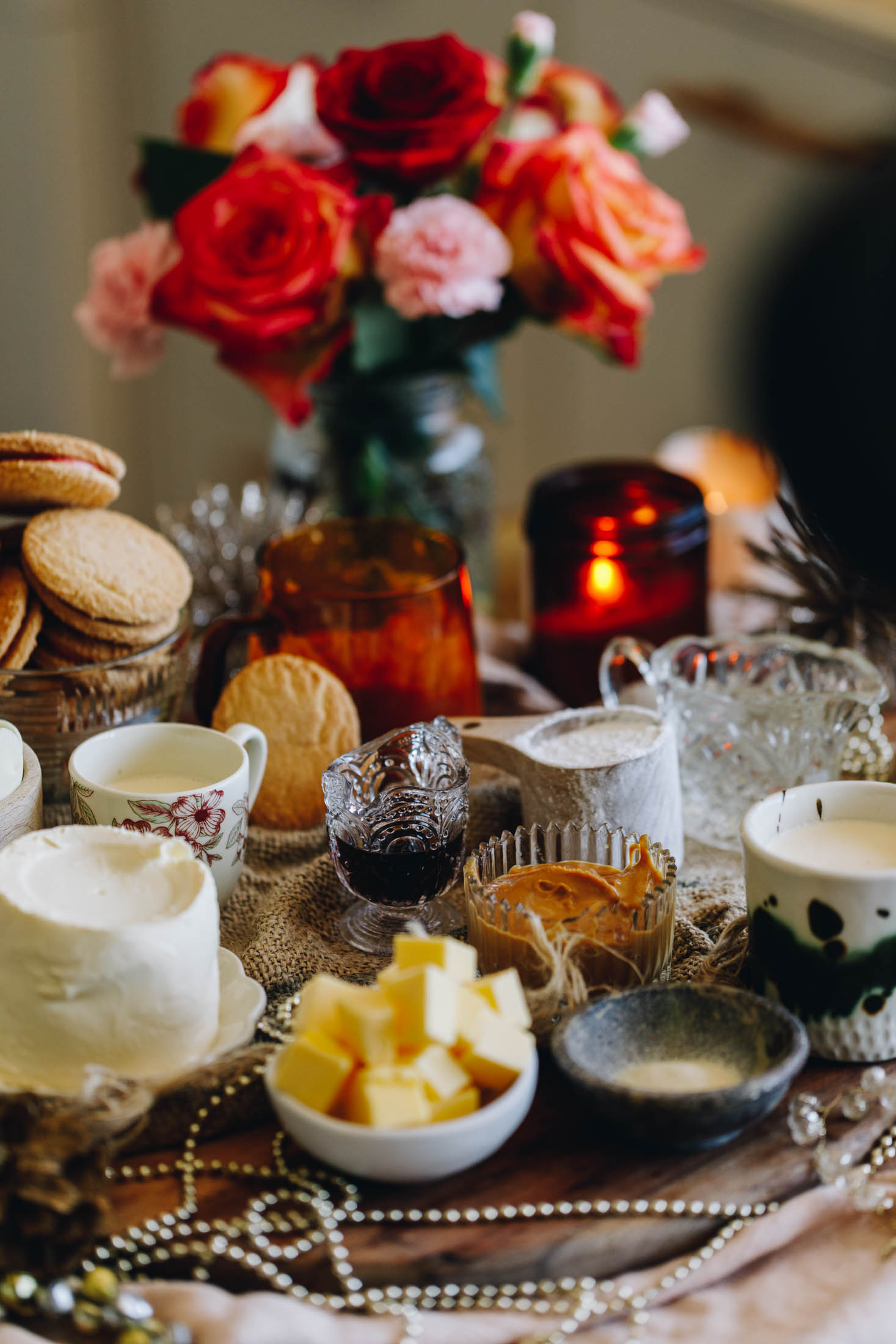 On a wooden board sits vintage bowls and small plates that have pan cotta ingredients on it. There is butter, cream cheese, peanut butter, biscuits and gelatine powder in view. They are sitting on a wooden table with natural fabrics on it, an orange candle and orange and pink flowers are in the background.