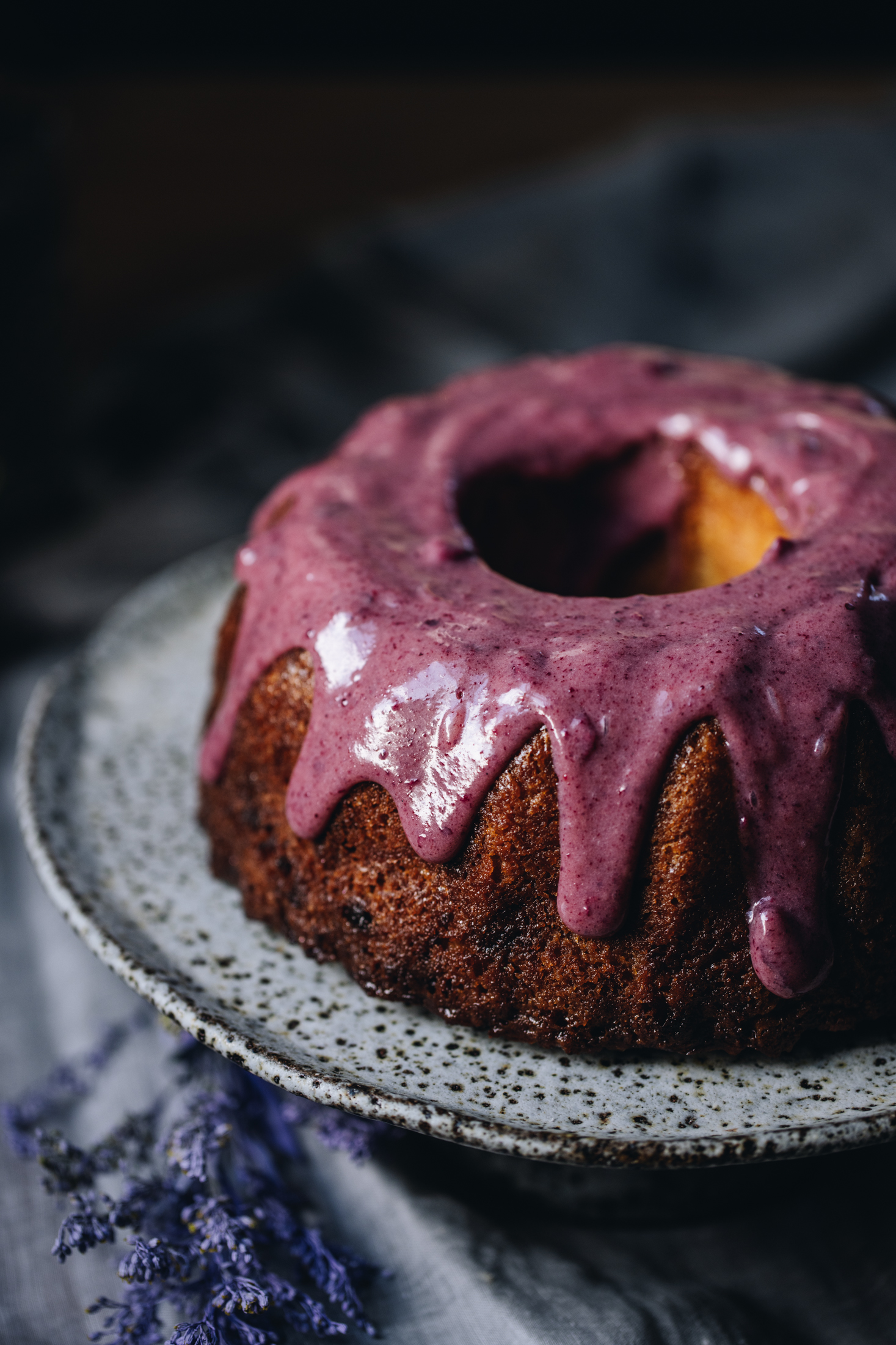 Sitting on natural fabric is a ceramic cake stand with a lemon syrup bundt cake on top of it. It is decorated with a white chocolate and plum ganache dripping down the sides of it. 
