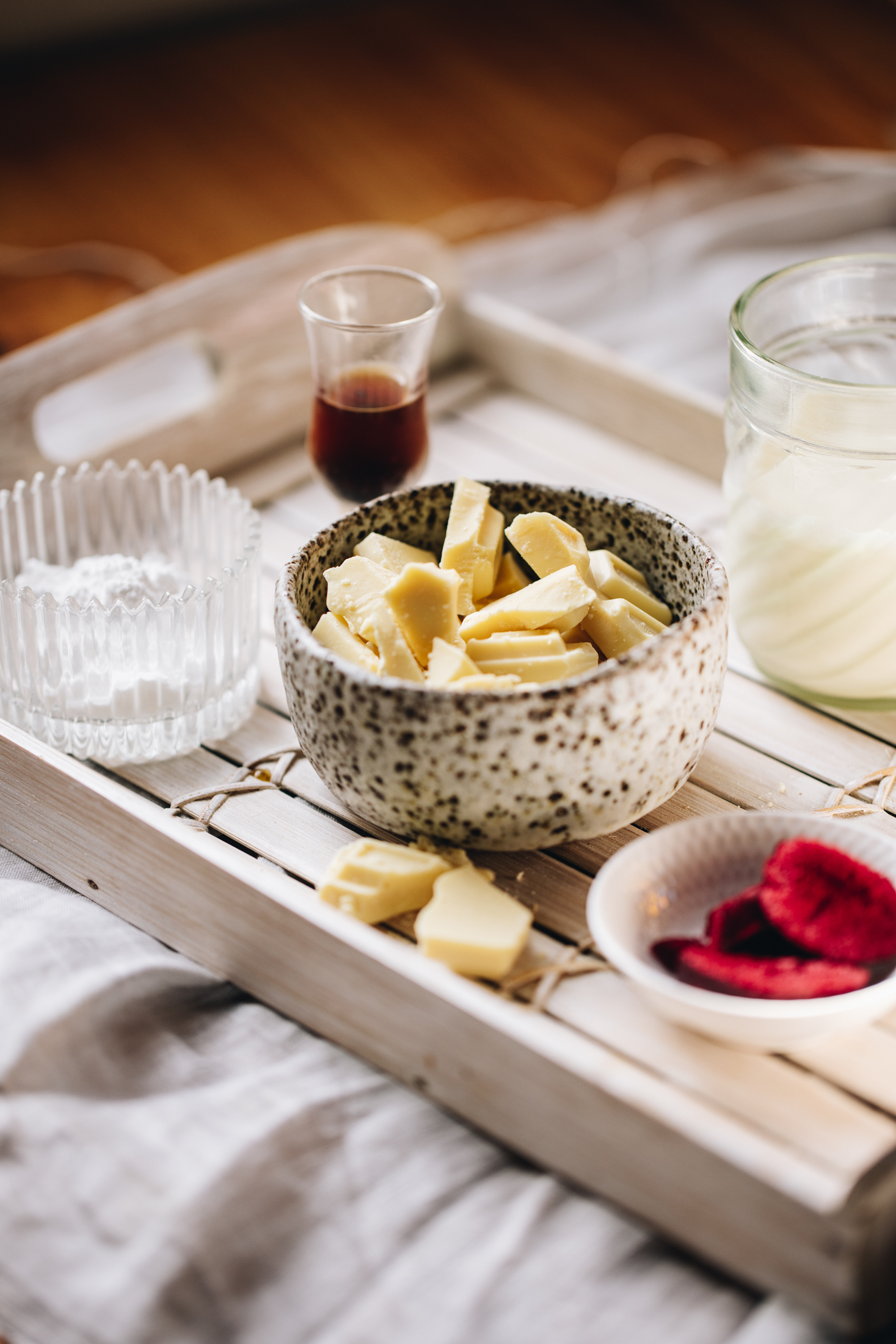 A white board is sitting on top of a light grey cloth. On it is various vintage bowls with ganache ingredients in it. There is white chocolate, freeze-dried plums and vanilla in view. 