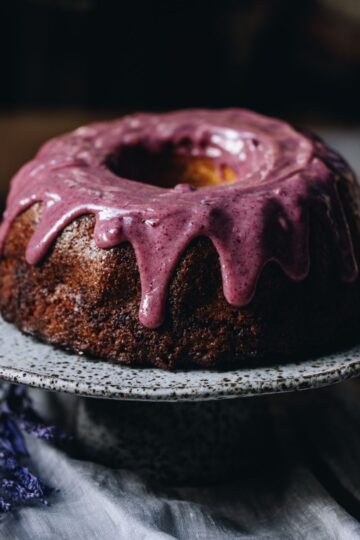 Sitting on natural fabric is a ceramic cake stand with a lemon syrup bundt cake on top of it. It is decorated with a white chocolate and plum ganache dripping down the sides of it. A purple flowers sits underneath it.