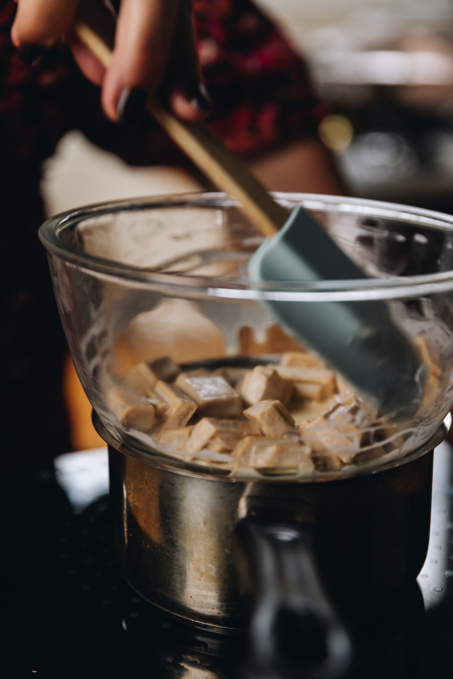 A silver pot sits on a a black portable stove with a glass bowl sitting snug on top of it. The bowl has white chocolate and cream in it and is being stirred by Naomi with a blue spatula with a wooden handle. 