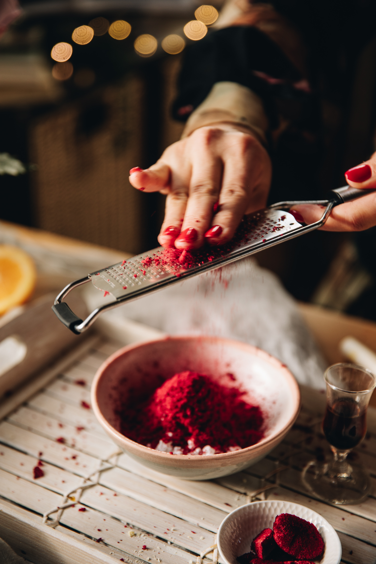 A small pink bowl sits on top of a white wooden board. In the bowl is freeze-dried plums that are being grated by Naomi using a fine grater.  Fairy lights are seen in the background. 