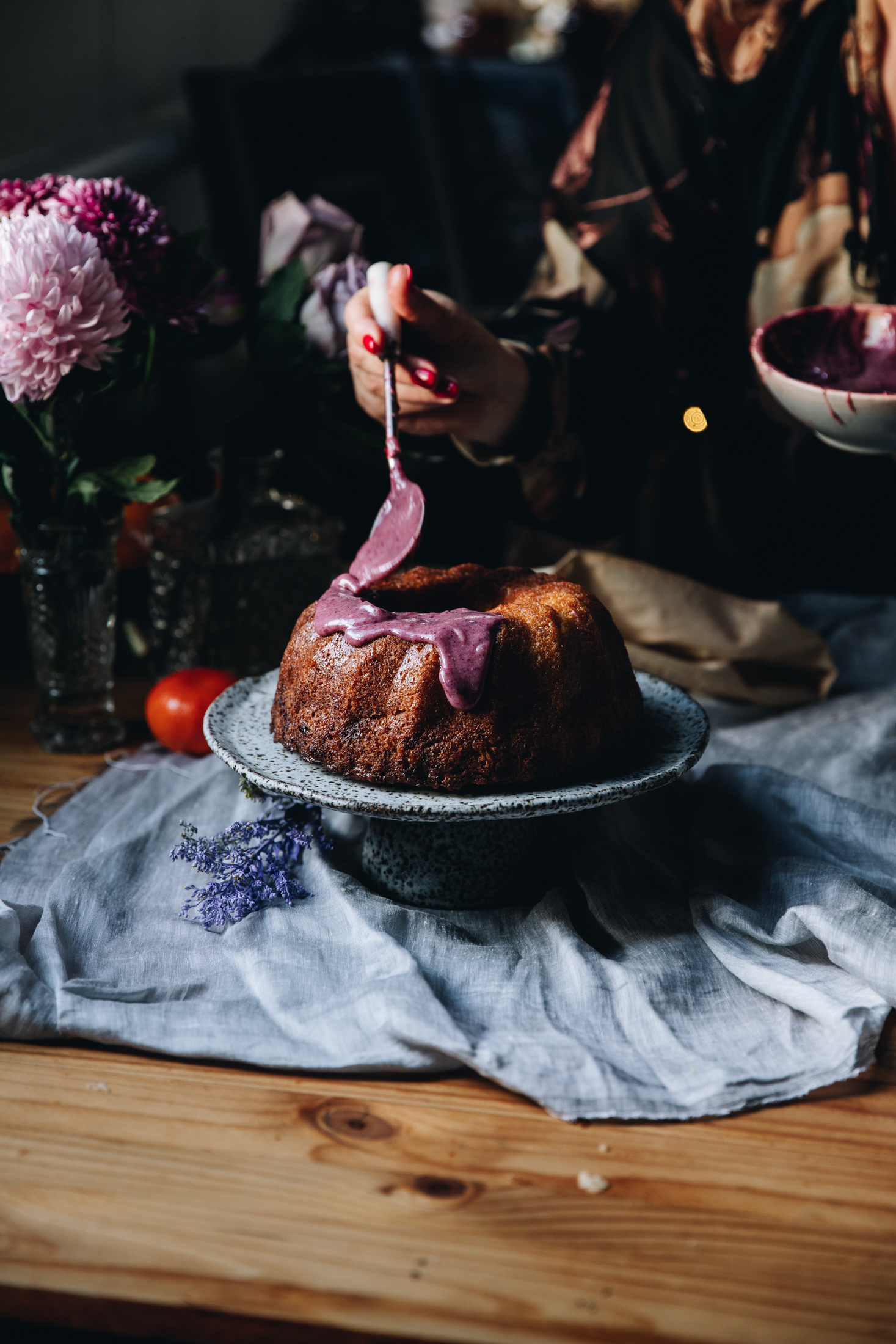 Sitting on natural fabric is a ceramic cake stand with a lemon syrup bundt cake on top of it. It is decorated with a white chocolate and plum ganache dripping down the sides of it. Naomi has a spoon and is adding more to the cake. A bunch of flowers sit in the background.