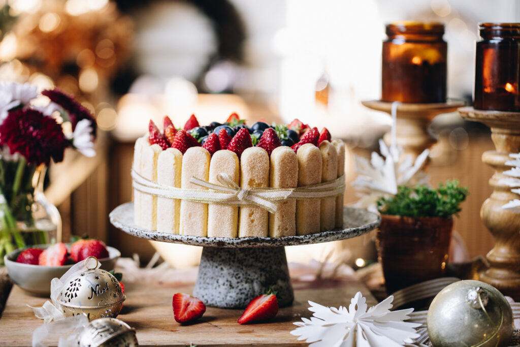 A no-bake Chocolate Charlotte Cake is decorated with berries. It is been tied with a gold ribbon, with a bow at the front. It is on a ceramic cake stand, on a wooden bench. It is surrounded by candles and Christmas decorations.