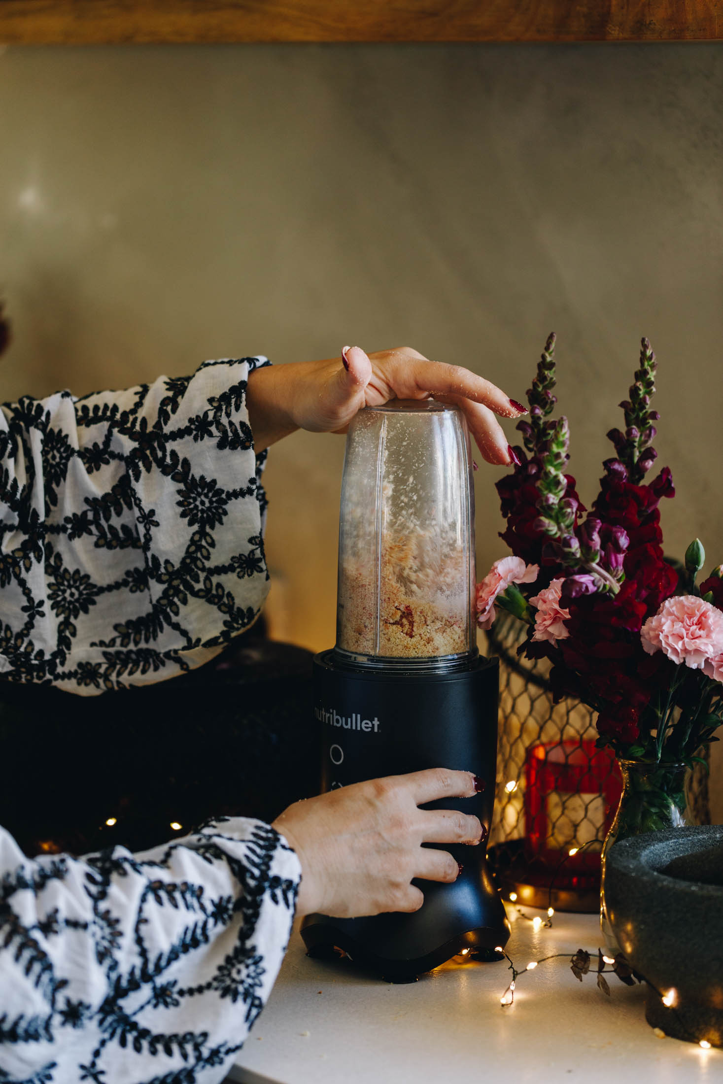 On a white stone bench is a small black blender with biscuit in it that are being crushed. Naomi is holding it with her hands. There are fairy lights and flowers in the background.