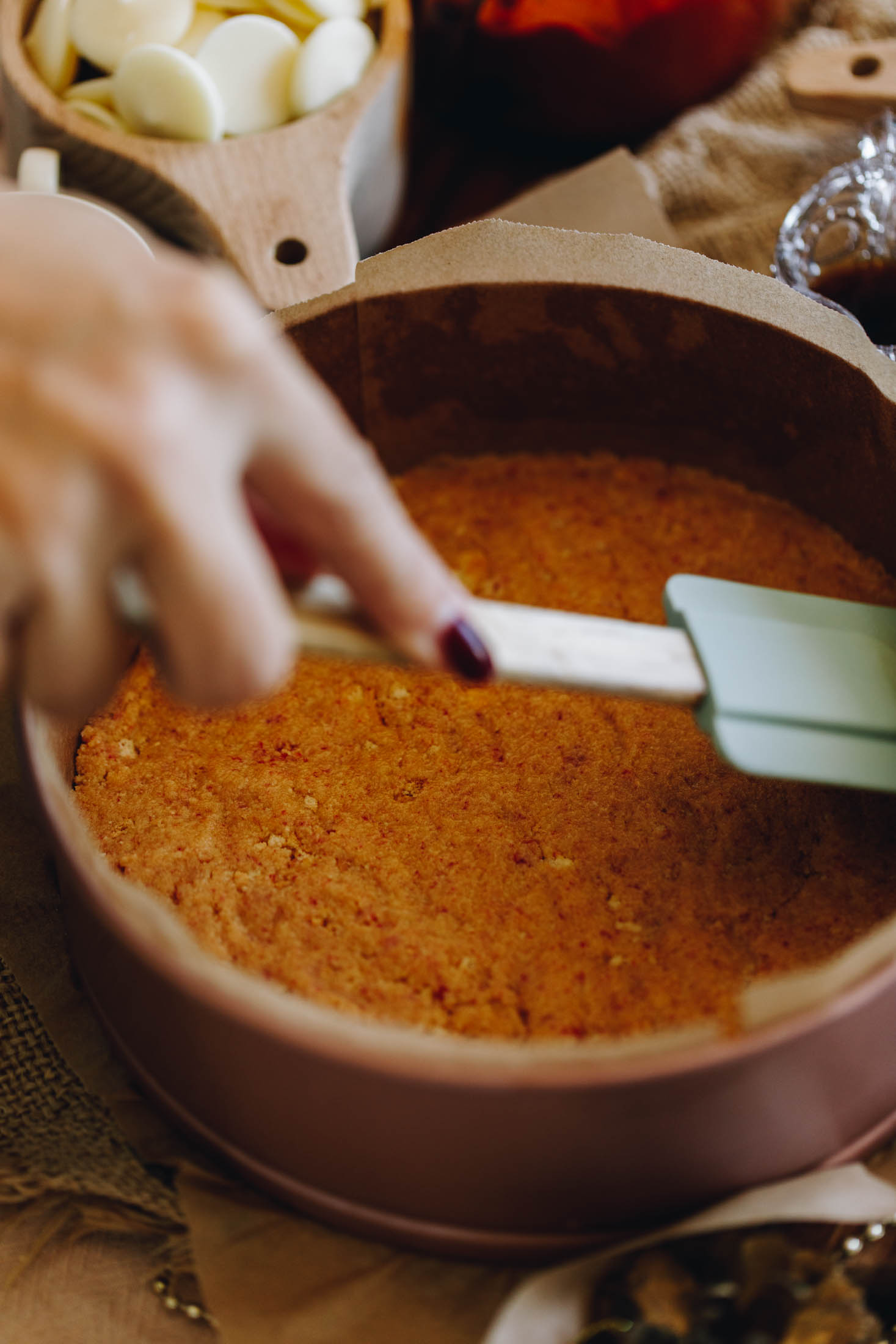 A rose gold cake tin lined with brown baking paper is on a table with natural fabric underneath it. In the cake tin is biscuit crumb that is being pressed down with a blue spatula with a wooden handle. White chocolate buttons in a wooden cup are in the background.
