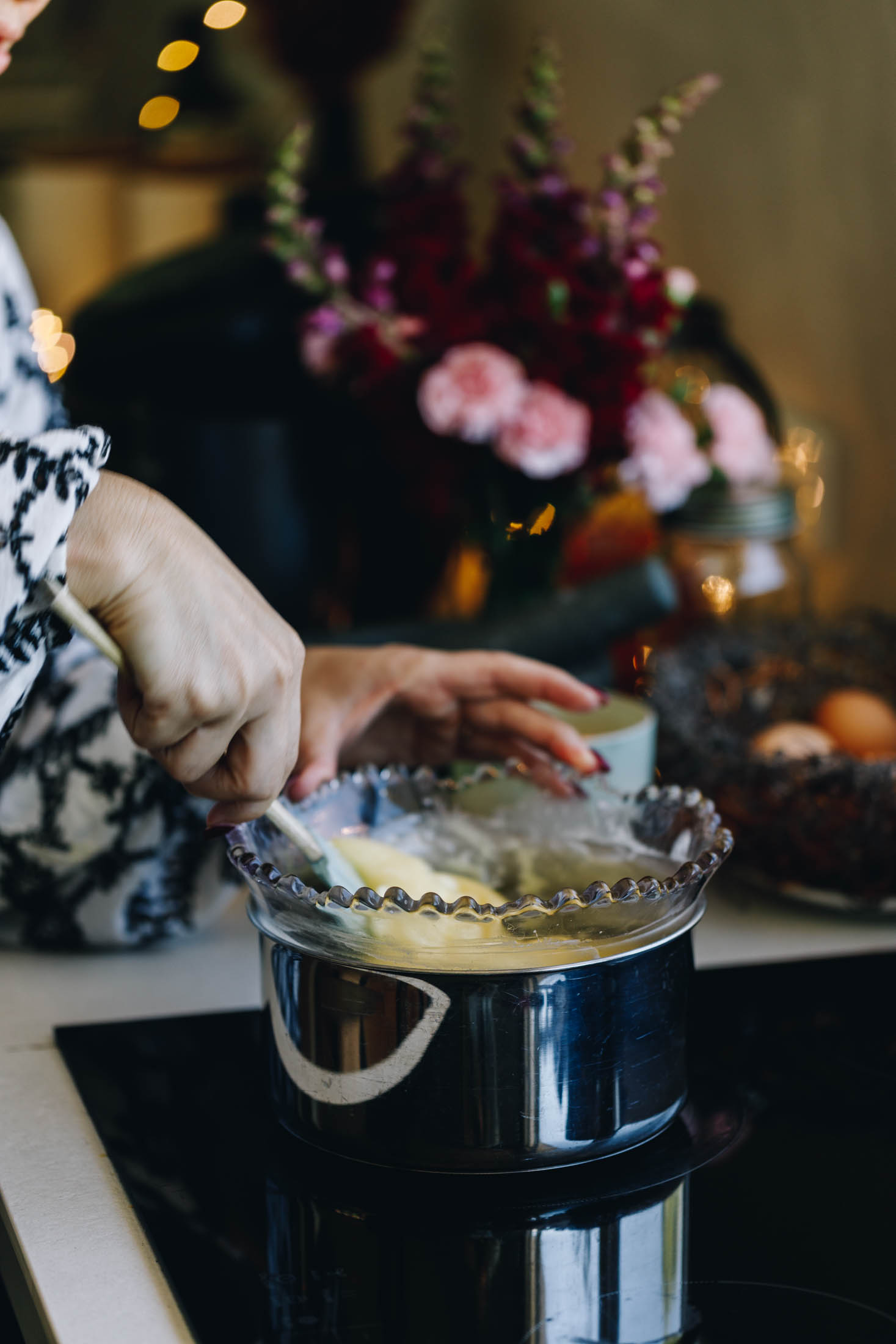 On a black stove sits a silver pot with a purple vintage bowl sitting inside it. It has a mixture of cream and white chocolate that has melted. It is being stirred with a blue spatula with a wooden handle. Flowers, fairy lights and a basket of eggs are in the background.