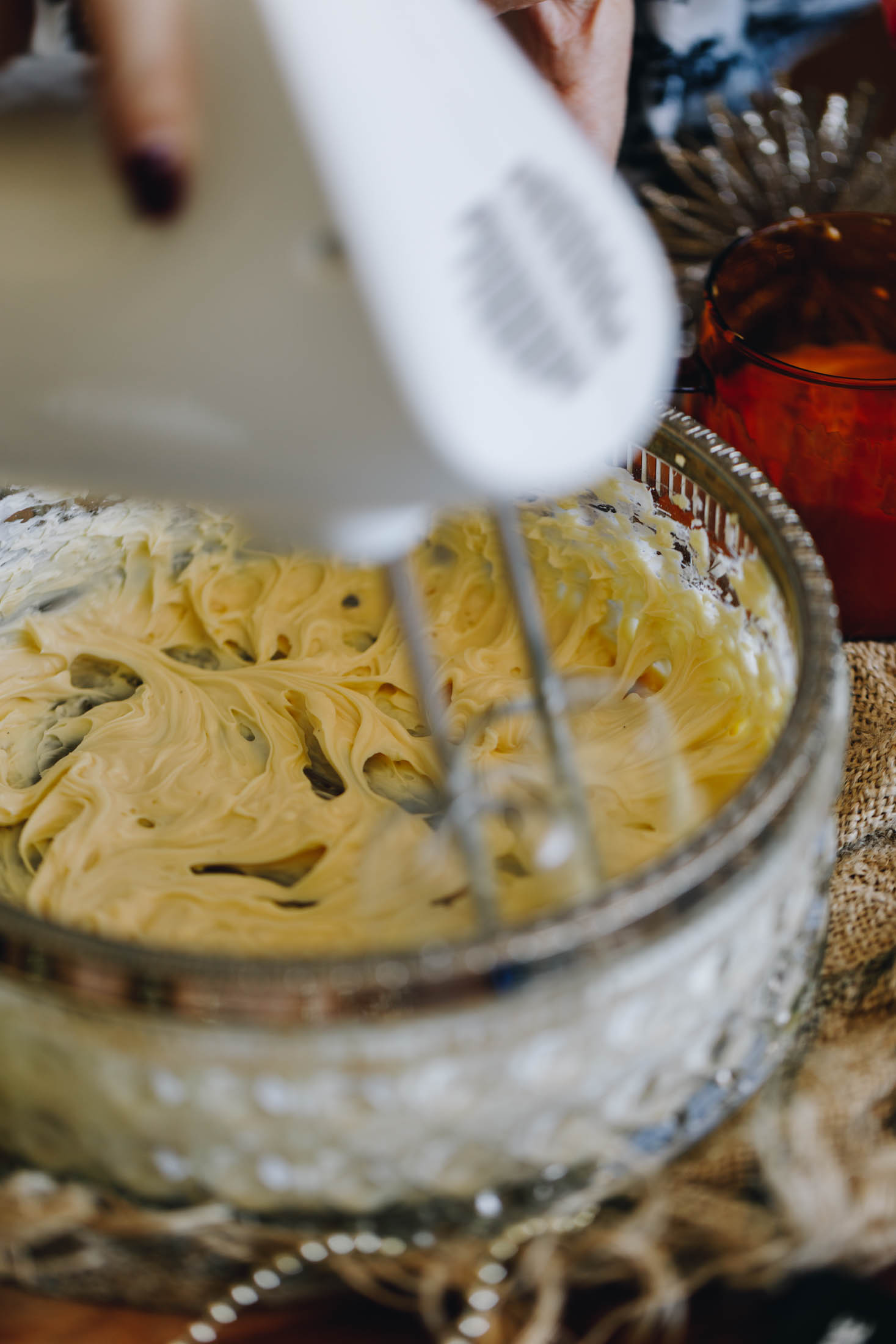 A vintage bowl with a silver trim sits on top of table with natural fabric on it. In the bowl cream cheese is being whipped by a white hand mixer. 