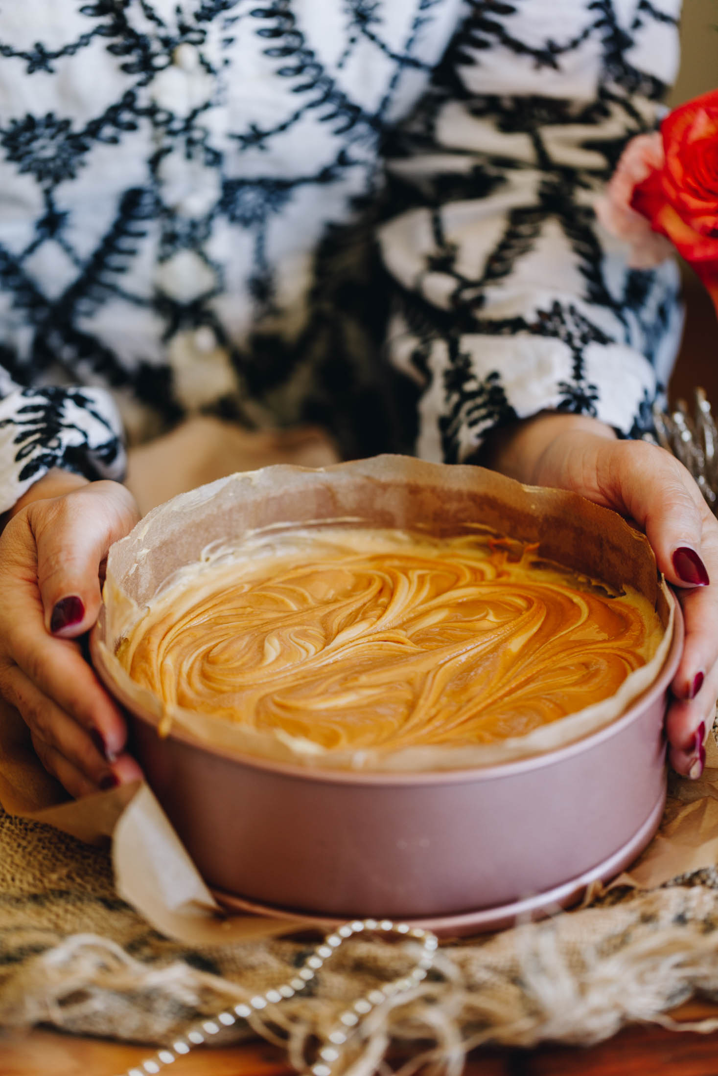 A rose gold cake tin lined with brown baking paper is on a table with natural fabric underneath it. In the cake tin is cheesecake mixture that has had peanut butter swirled in to it. Naomi is holding either side of the cake tin with her hands ready to remove it. 