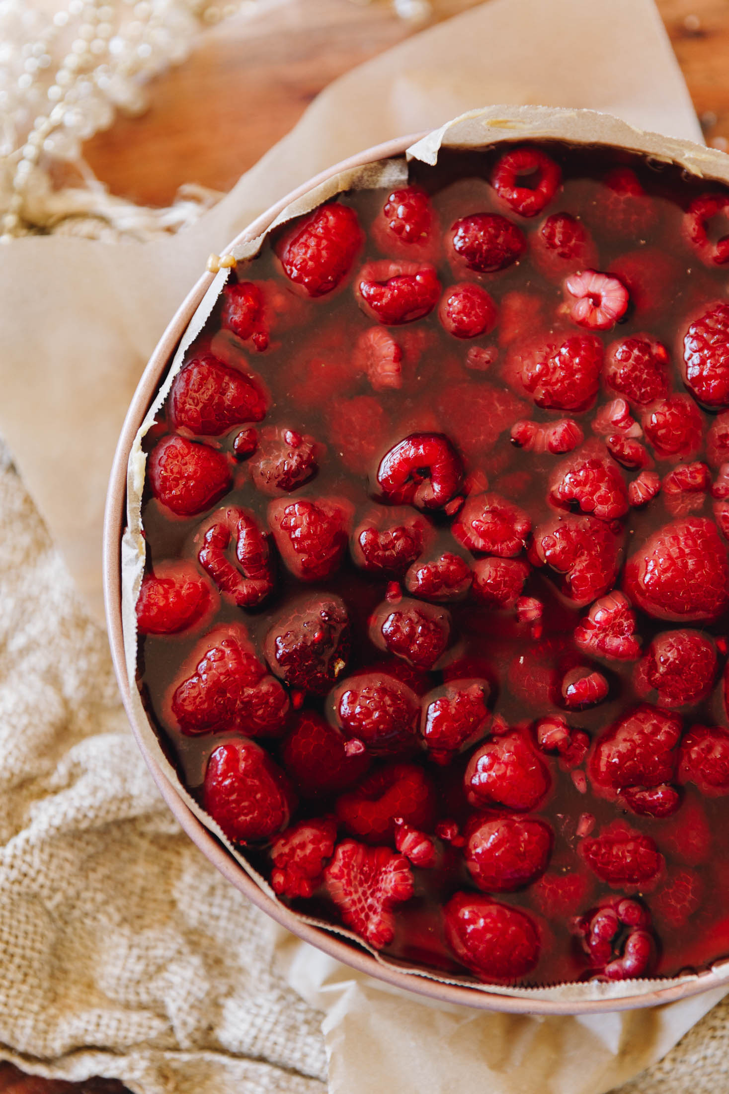 A top shot shows a rose gold cake tin lined with brown baking paper on a table with natural fabric underneath it. In the cake tin is the peanut butter cheesecake with raspberries and a pomegranate jelly on it. 