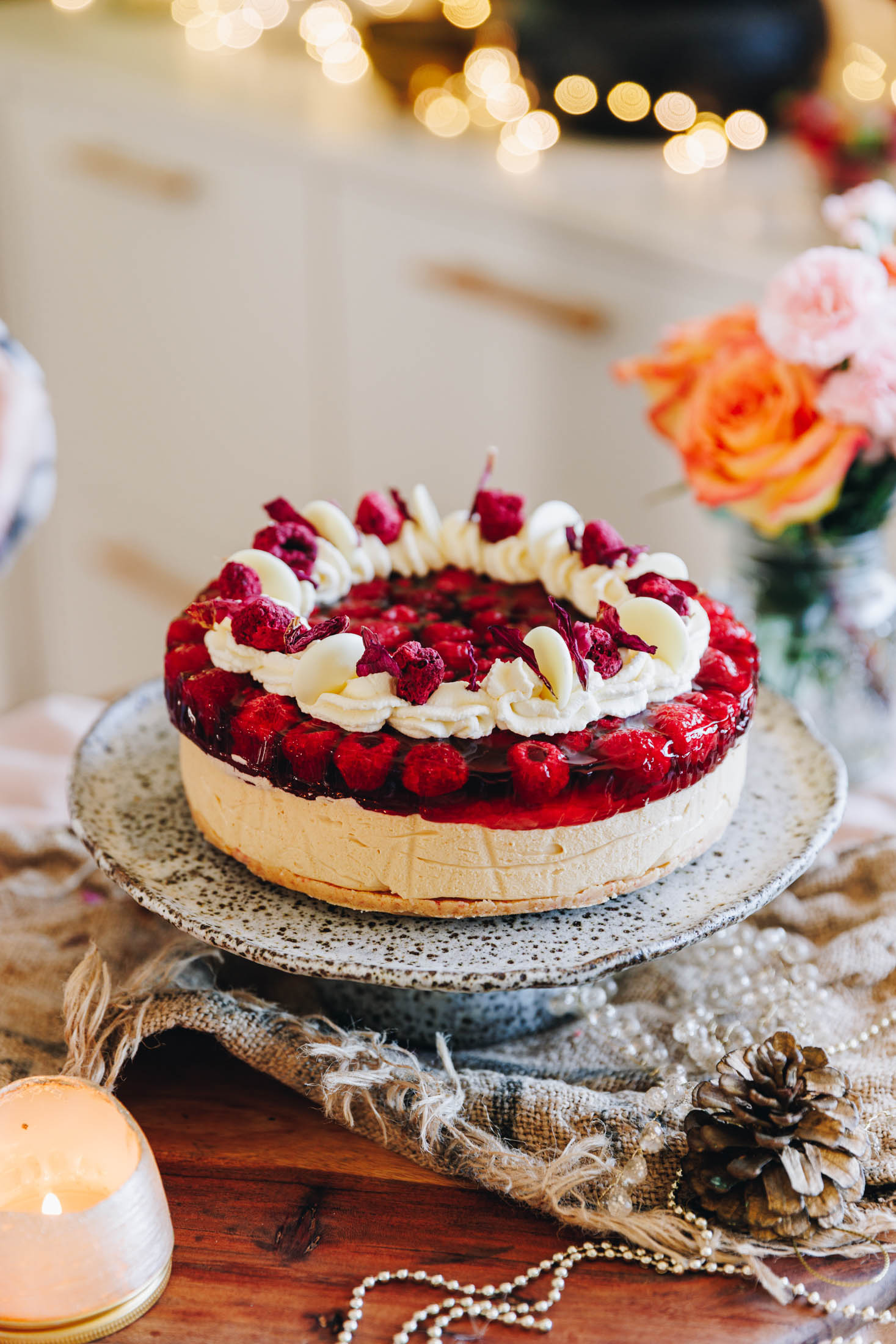 The peanut butter jelly cheesecake has been decorated with piped cream, freeze-dried raspberries and flower petals. It is on a ceramic cake stand which is sitting on a wooden board with natural fabrics and Christmas decorations. 