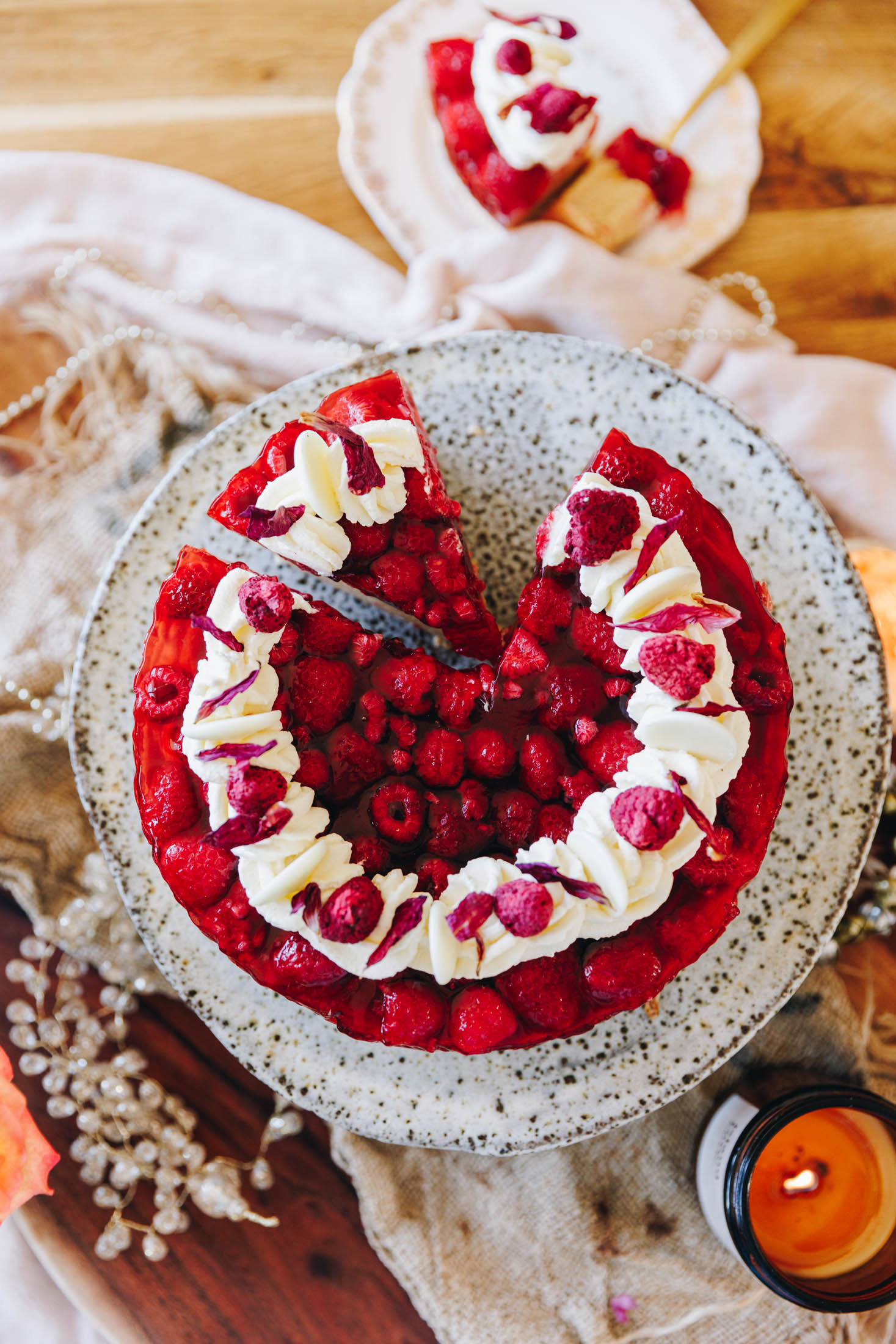 A top shot shows the peanut butter jelly cheesecake that has been decorated with piped cream, freeze-dried raspberries and flower petals. It is on a ceramic cake stand which is sitting on a wooden board with natural fabrics and Christmas decorations. There is a slice removed that sits on a white plate and a fork. Another piece has been cut on the cake stand too. An amber candle burns in the corner.