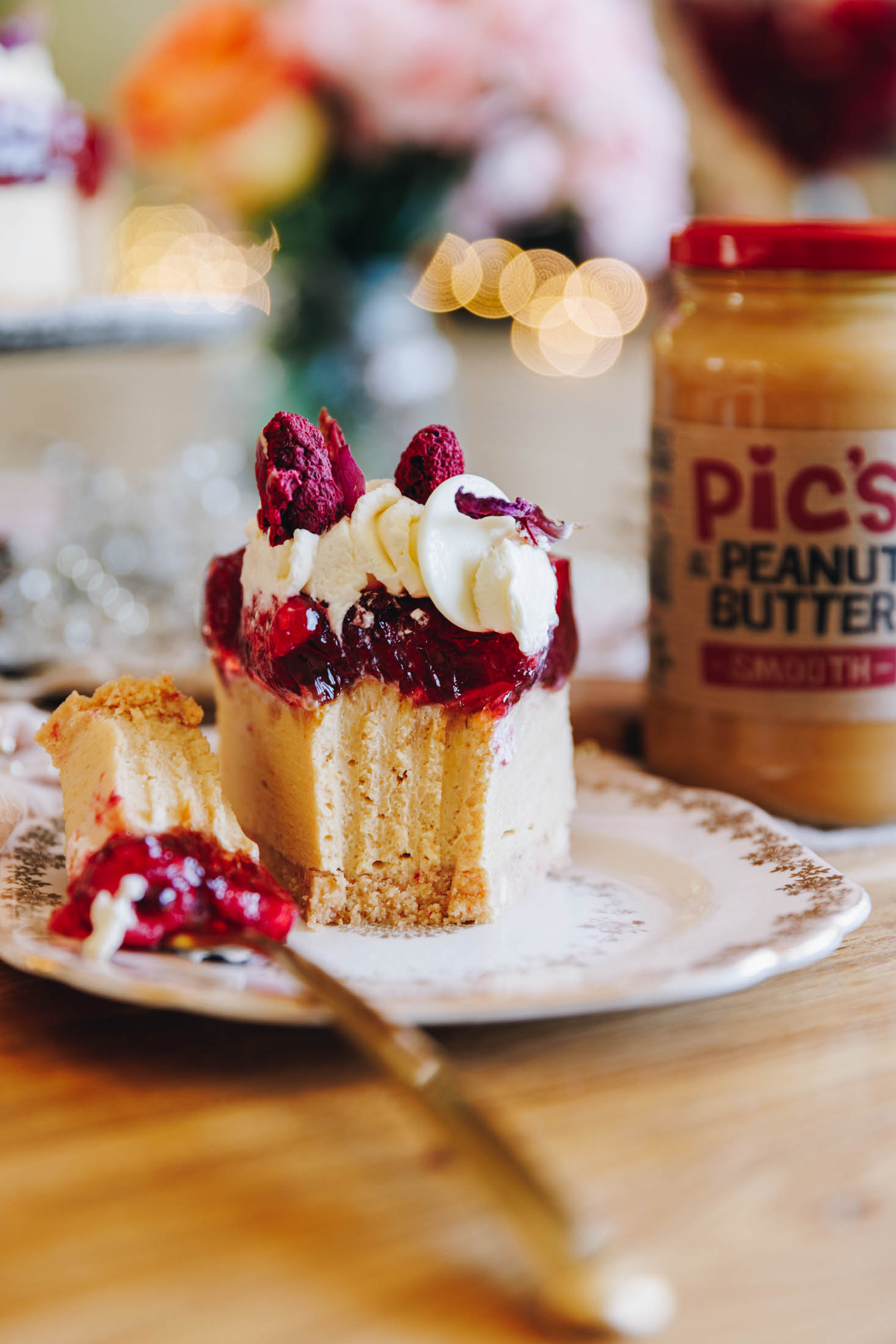 A single slice of peanut butter jelly cheesecake sits on a pink plate with a vintage frill. It has been cut into with a fork and the golden fork still has the remnants on it. A jar of PIc's Smooth peanut butter sits behind it with blurred flowers and fairy lights in the background. 