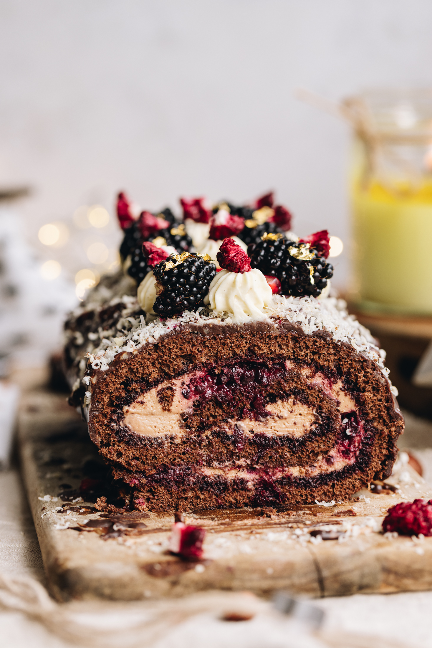 A chocolate sponge roll is on top of a wooden board that has freeze-dried boysenberries sprinkled at the front of the board. The sponge has been cut open and reveals the chocolate cream and berries that are inside the sponge. Chocolate covers the roll and it is dusted with grated hazelnuts. Piped cream is piped on top of the sponge roll, and pieces of freeze-dried boysenberries on top of each one. Blackberries and gold leaf are on top also. A yellow candle burns in the background along with golden fairy lights. 