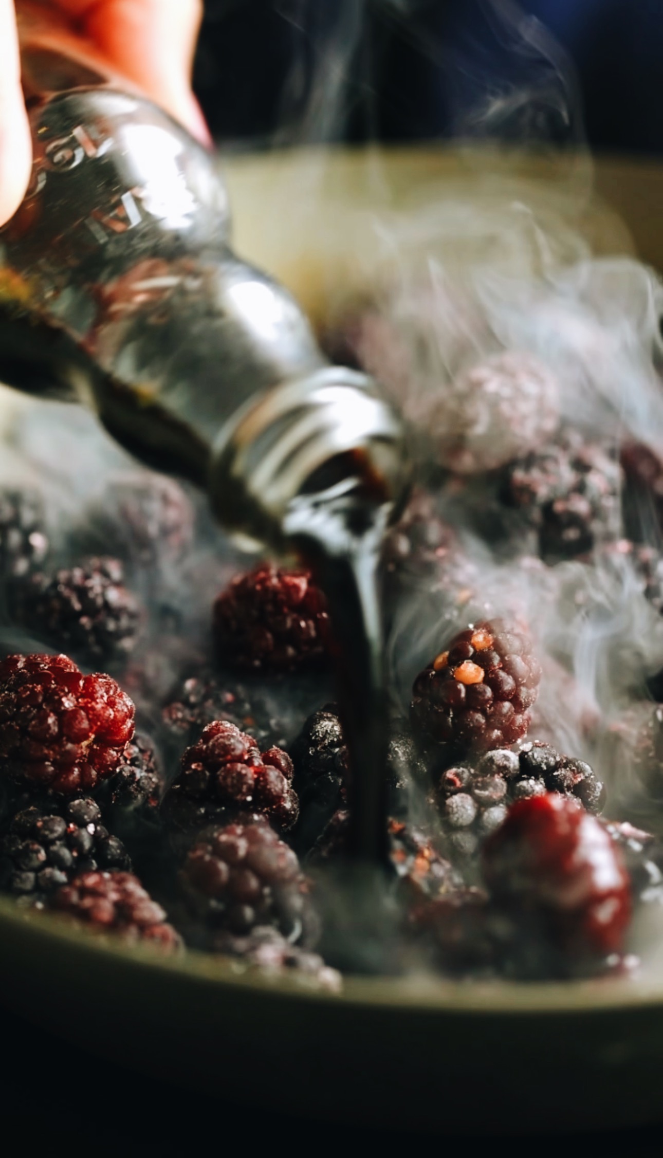 A close up shot shows a frying pan with blackberries in with steam rising off it. A vintage glass bottle is pouring in balsamic vinegar in to the berries. 