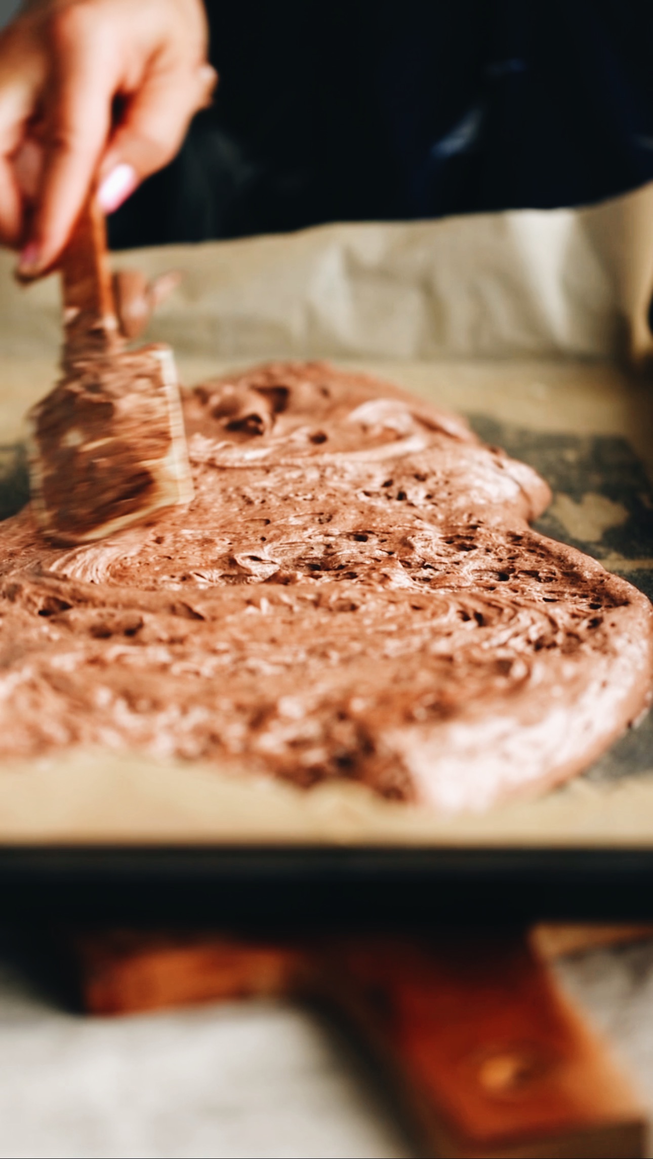 A black tray lined with brown baking paper is on a wooden chopping board. In the tray is chocolate sponge mixture that is being smoothed out with a spatula. 