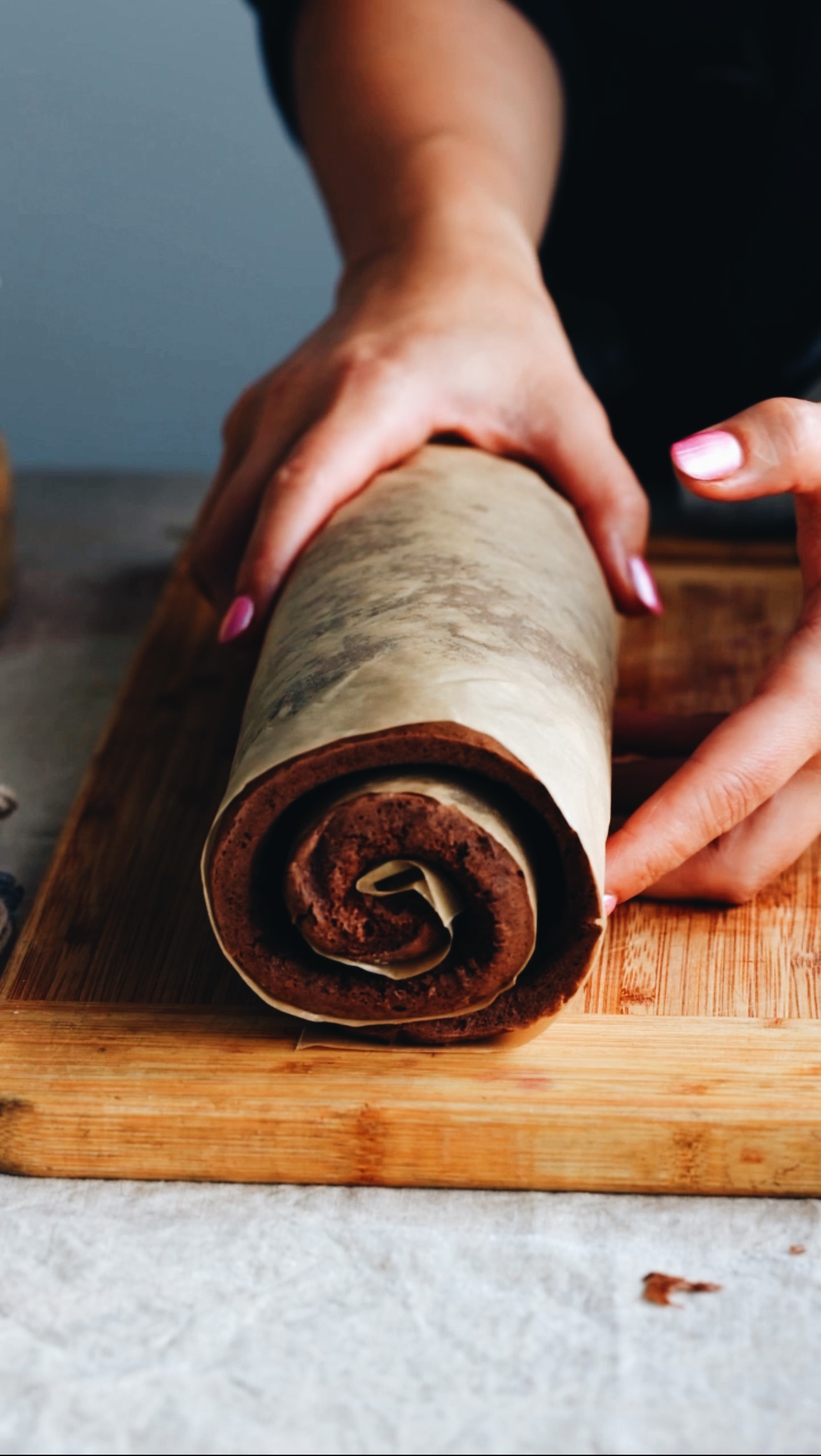 A baked sponge is rolled in brown baking paper and is facing the camera to show the spiral effect. It is on a wooden board on top of a grey table. It is being held by Naomi's hands. 