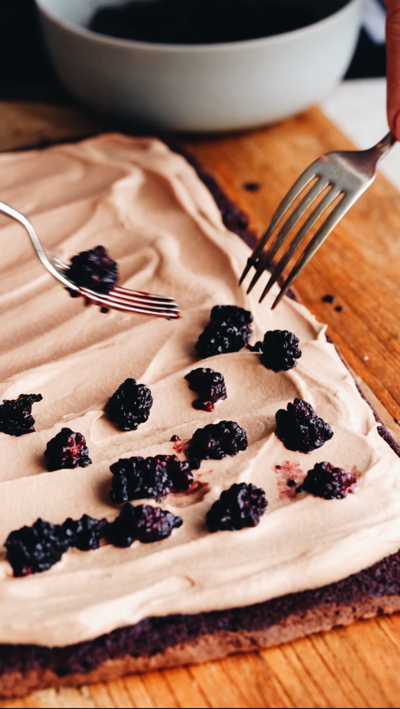 The chocolate sponge roll has been rolled out and is covered in chocolate cream and two forks are placing the cooked blackberries on to the stone. A grey bowl is in the background. The sponge is on a wooden board. 