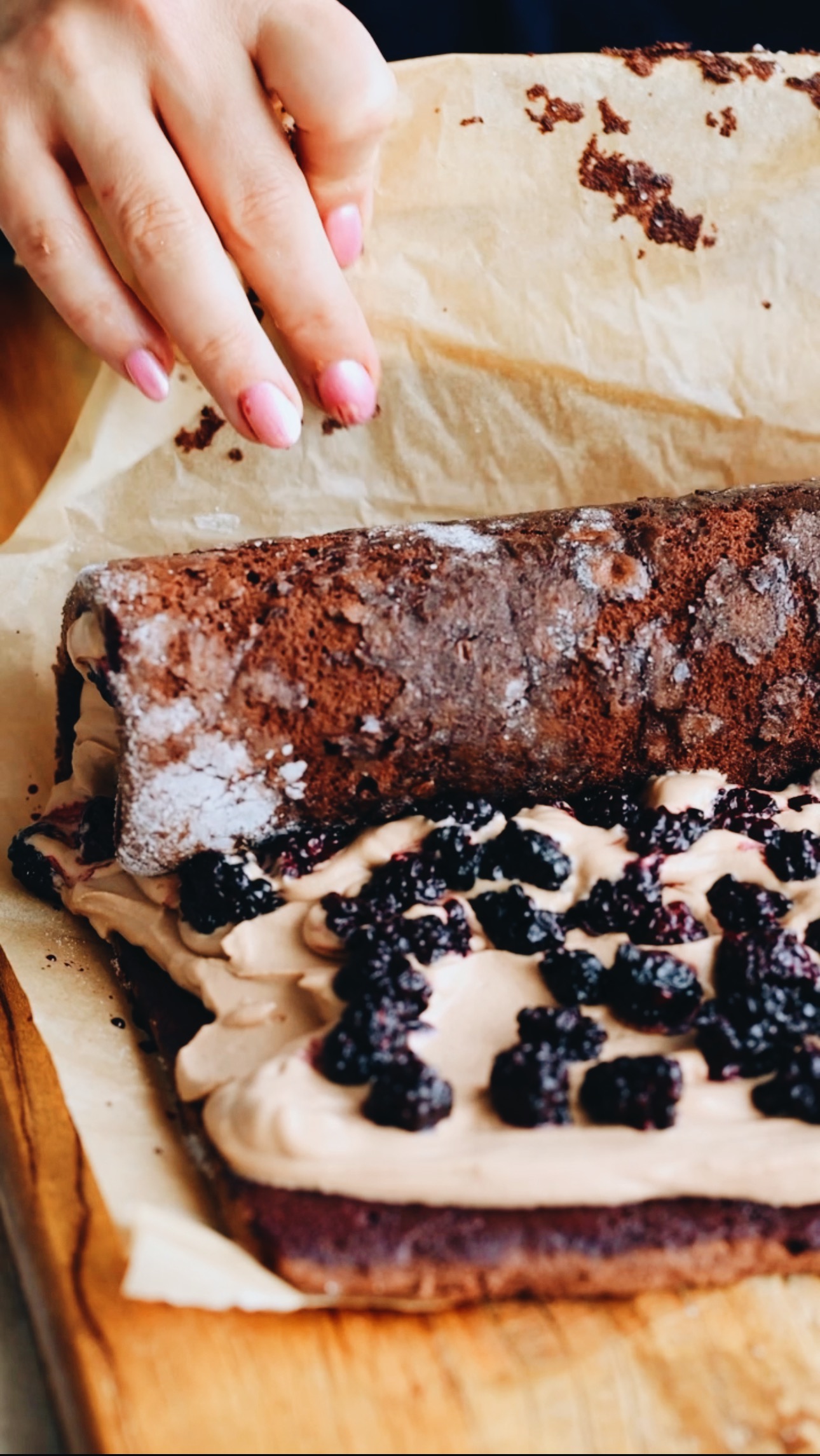 A chocolate sponge is being roll with chocolate cream and blackberries on it. Brown baking paper is being held by Naomi and she is using it to push the sponge forward. 