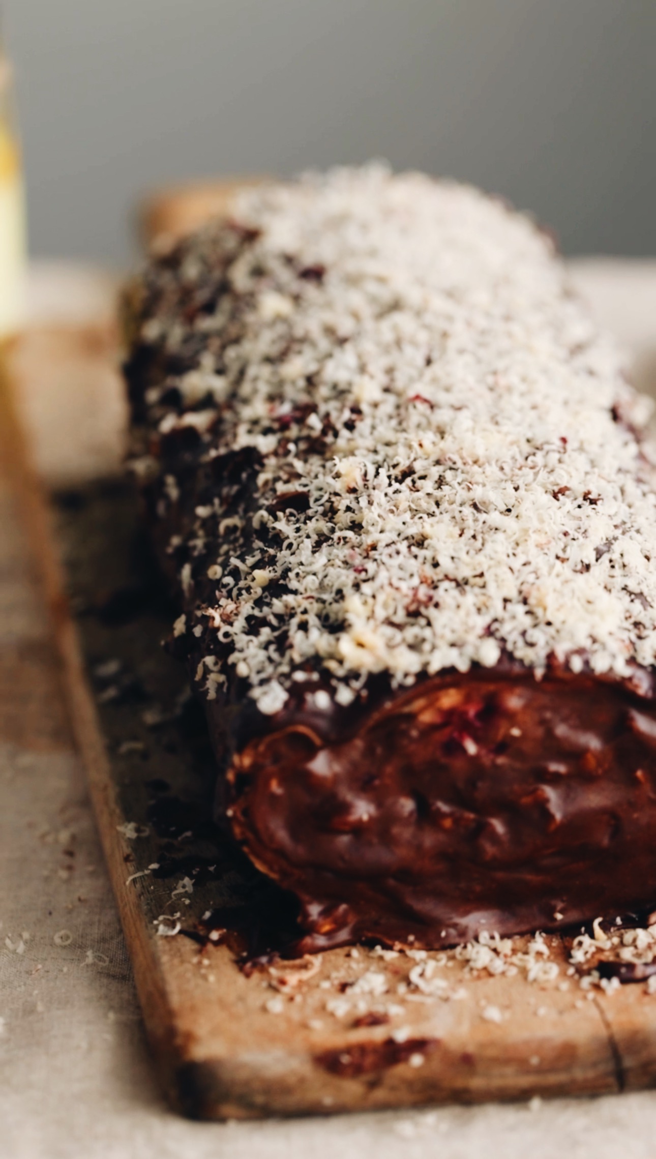 A chocolate sponge roll is sitting on a wooden board. It has been covered completely in chocolate and has grated hazelnut over the top of it that looks like snow. 