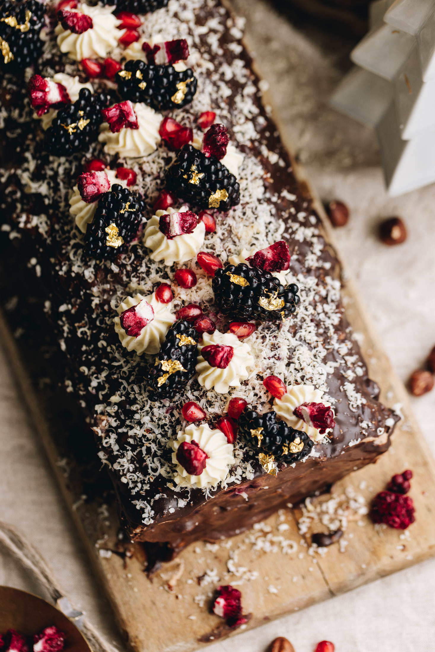 A top shot shows chocolate the covers the roll and it is dusted with grated hazelnuts. Piped cream is piped on top of the sponge roll, and pieces of freeze-dried boysenberries on top of each one. Blackberries and gold leaf are on top also.  There is a ceramic white Christmas tree in the corner of the shot. 