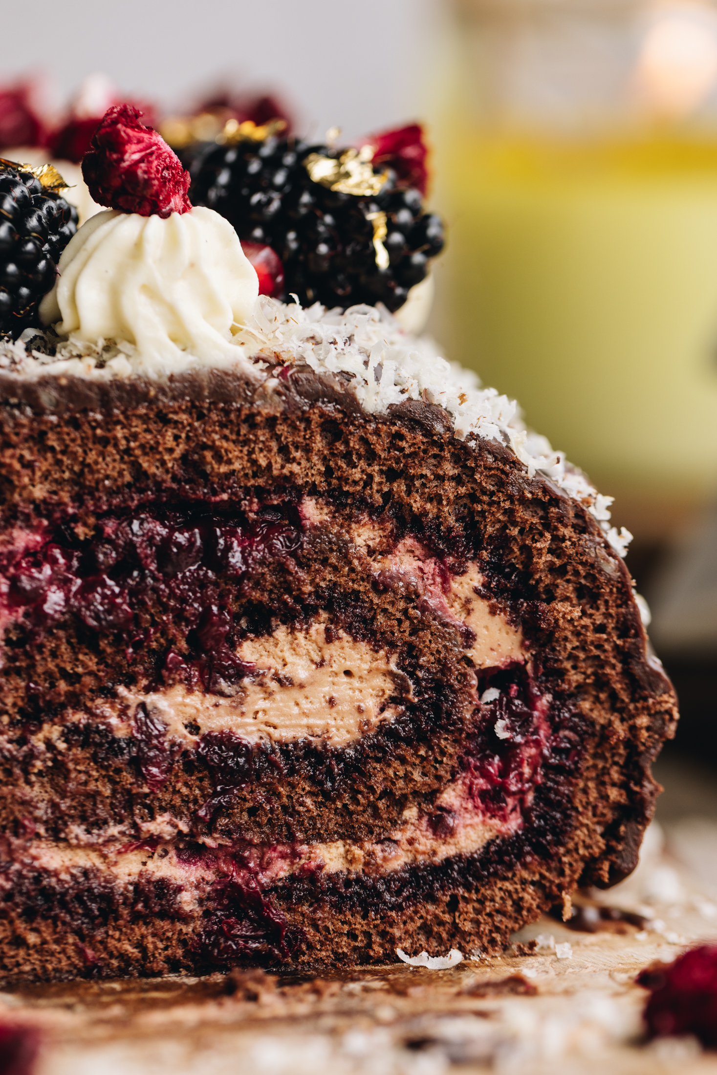 A close shot shows a chocolate sponge roll is on top of a wooden board that has freeze-dried boysenberries sprinkled at the front of the board. The sponge has been cut open and reveals the chocolate cream and berries that are inside the sponge. Chocolate covers the roll and it is dusted with grated hazelnuts. Piped cream is piped on top of the sponge roll, and pieces of freeze-dried boysenberries on top of each one. Blackberries and gold leaf are on top also. 