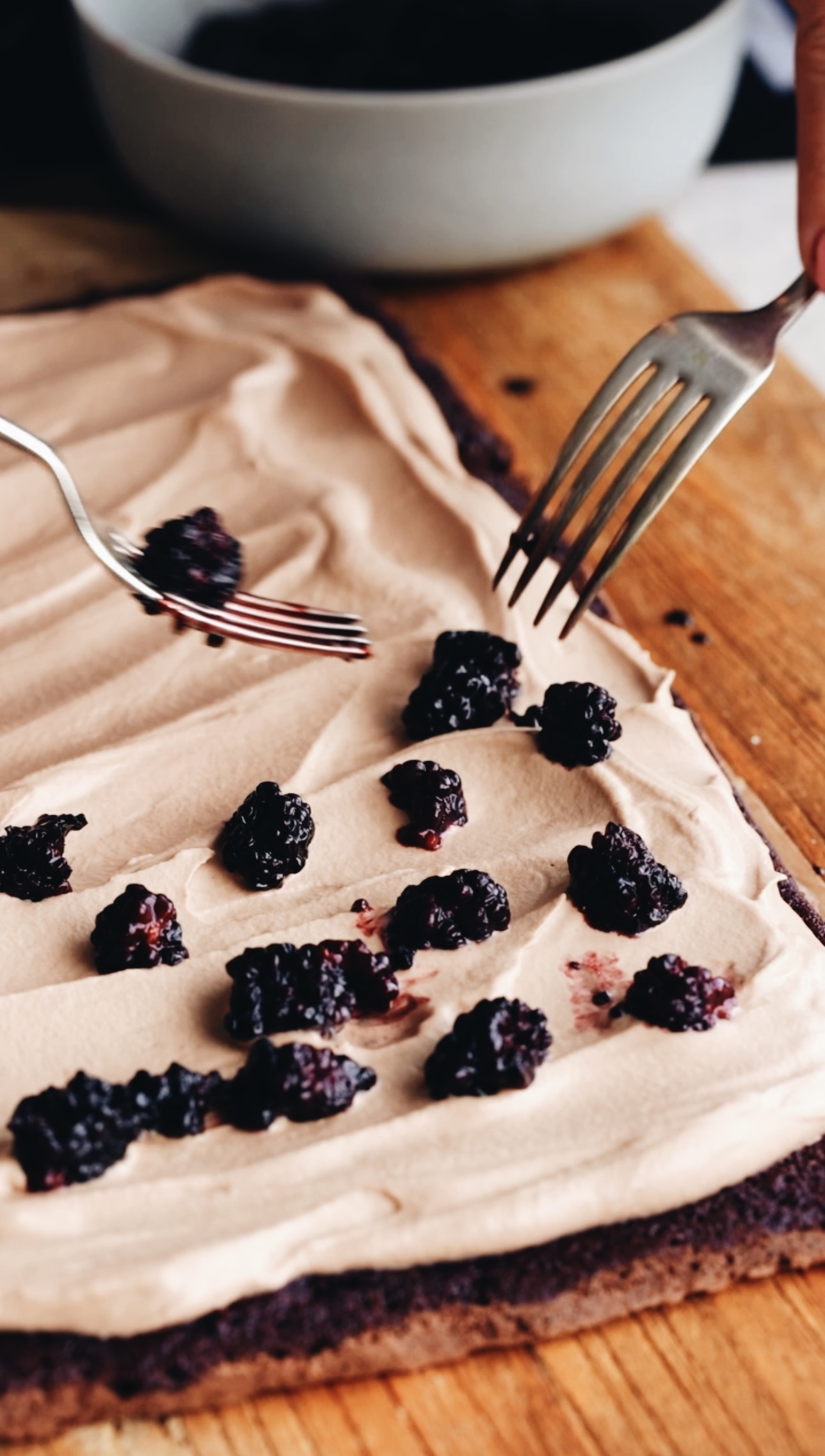 The chocolate sponge roll has been rolled out and is covered in chocolate cream and two forks are placing the cooked blackberries on to the stone. A grey bowl is in the background. The sponge is on a wooden board.
