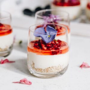 A decorated white chocolate mousse in a glass sits on a white table. In the glass is a biscuit base, a white chocolate mousse and finely chopped berries with juice on it and an edible purple flower on top. Half of another set mousse is behind it. In the far background is a white tray with more desserts in glasses.